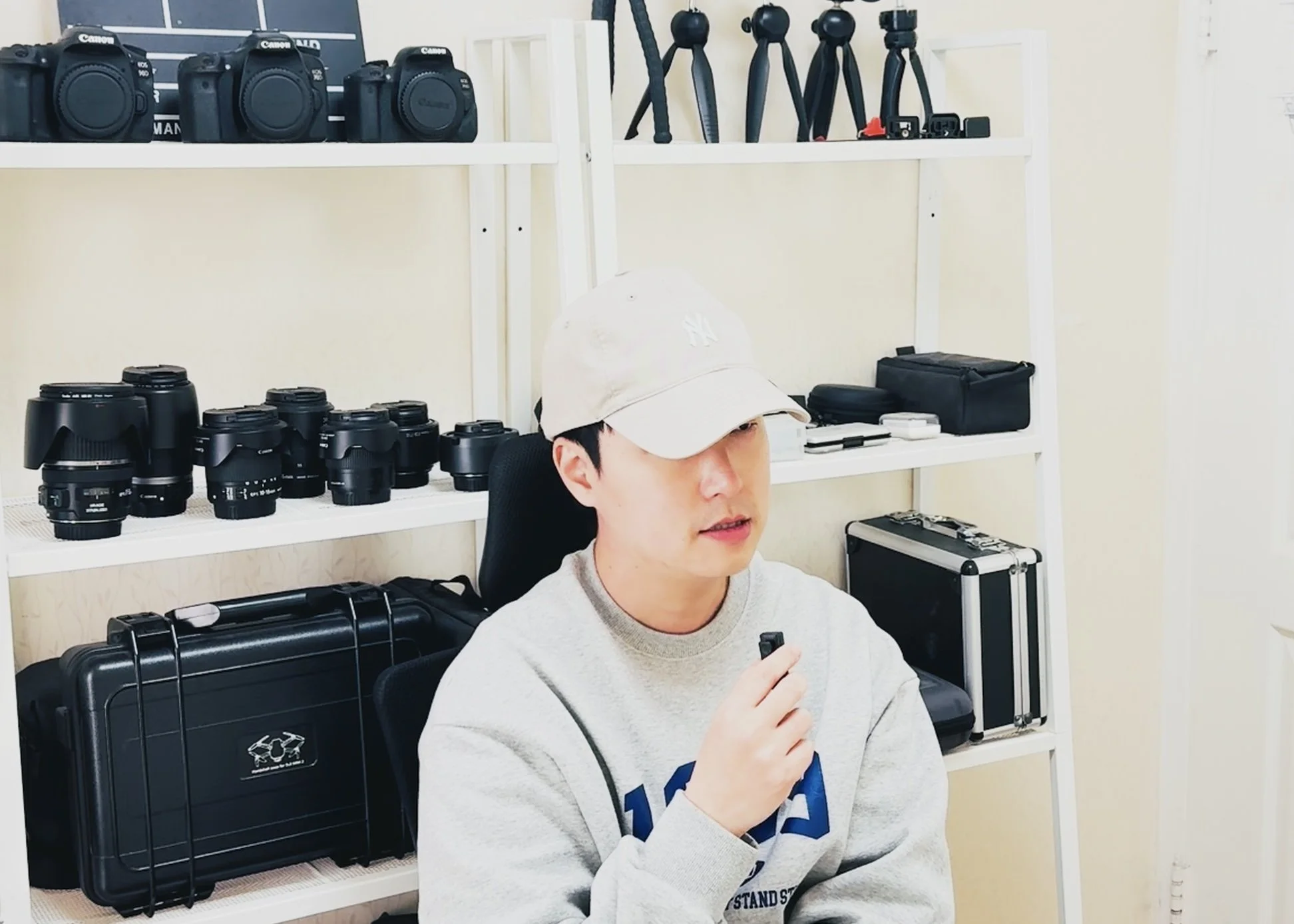 사진은 흰색 모자를 쓴 남자가 회의실에 앉아 있으며, 뒤에는 카메라와 렌즈, 삼각대, 기타 장비가 진열된 선반이 있다.