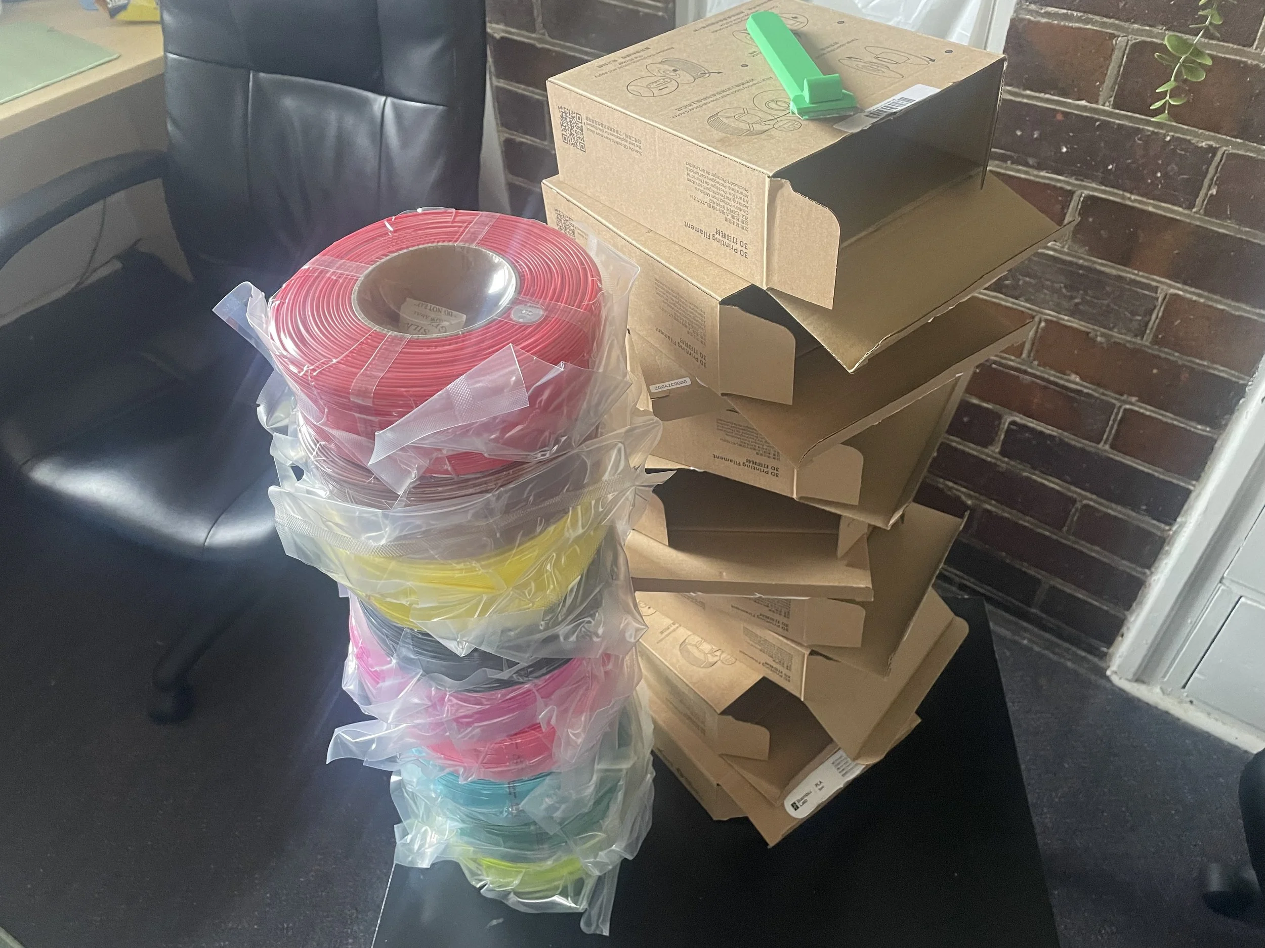 Stacked cardboard boxes on a table with a black office chair nearby, and a large roll of red and yellow plastic film wrapped in plastic in the foreground, against a brick wall background.