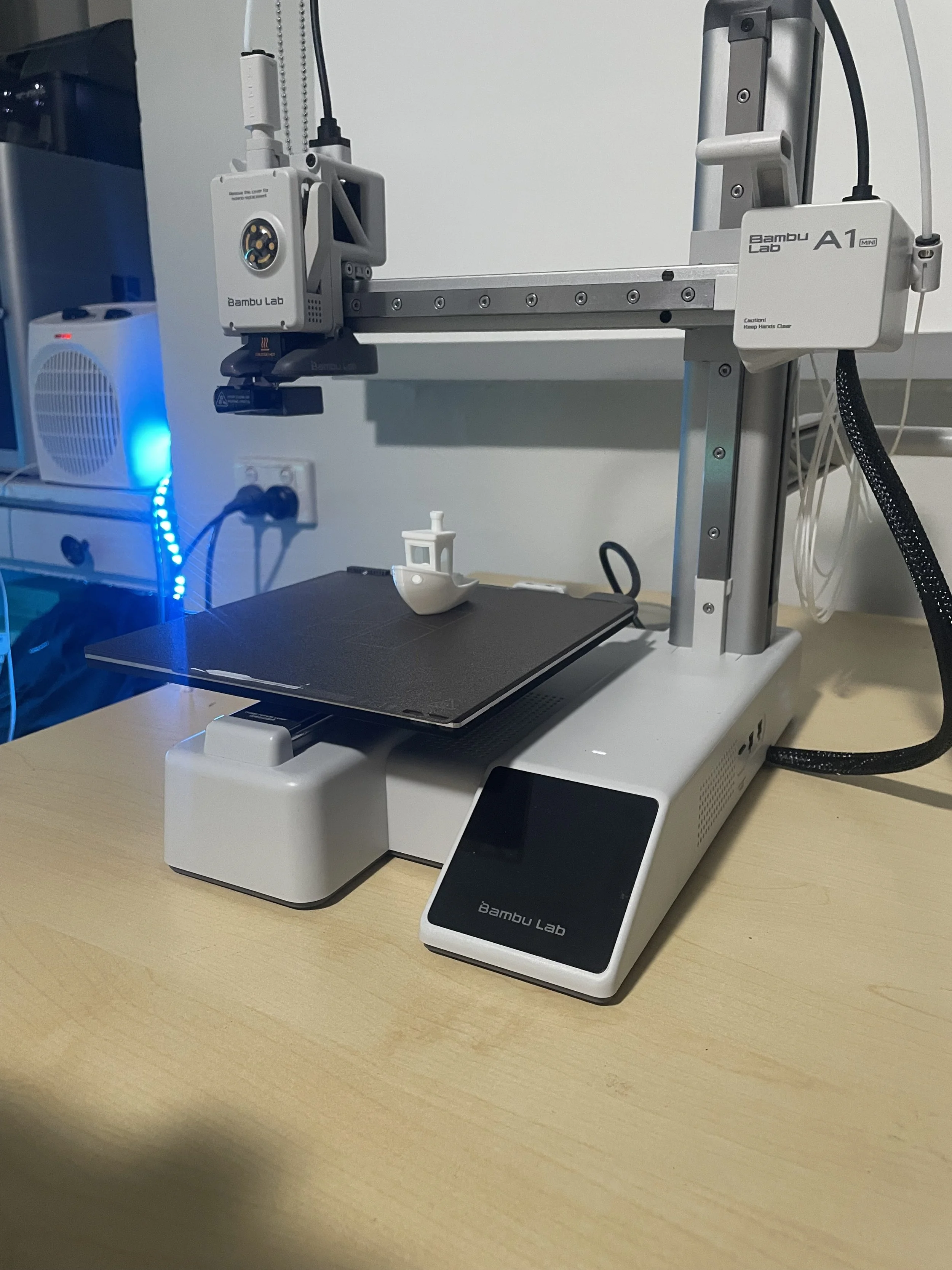 A 3D printer on a wooden desk with a small white boat model being printed. The printer has a touch screen and branding 'Bambu Lab'.