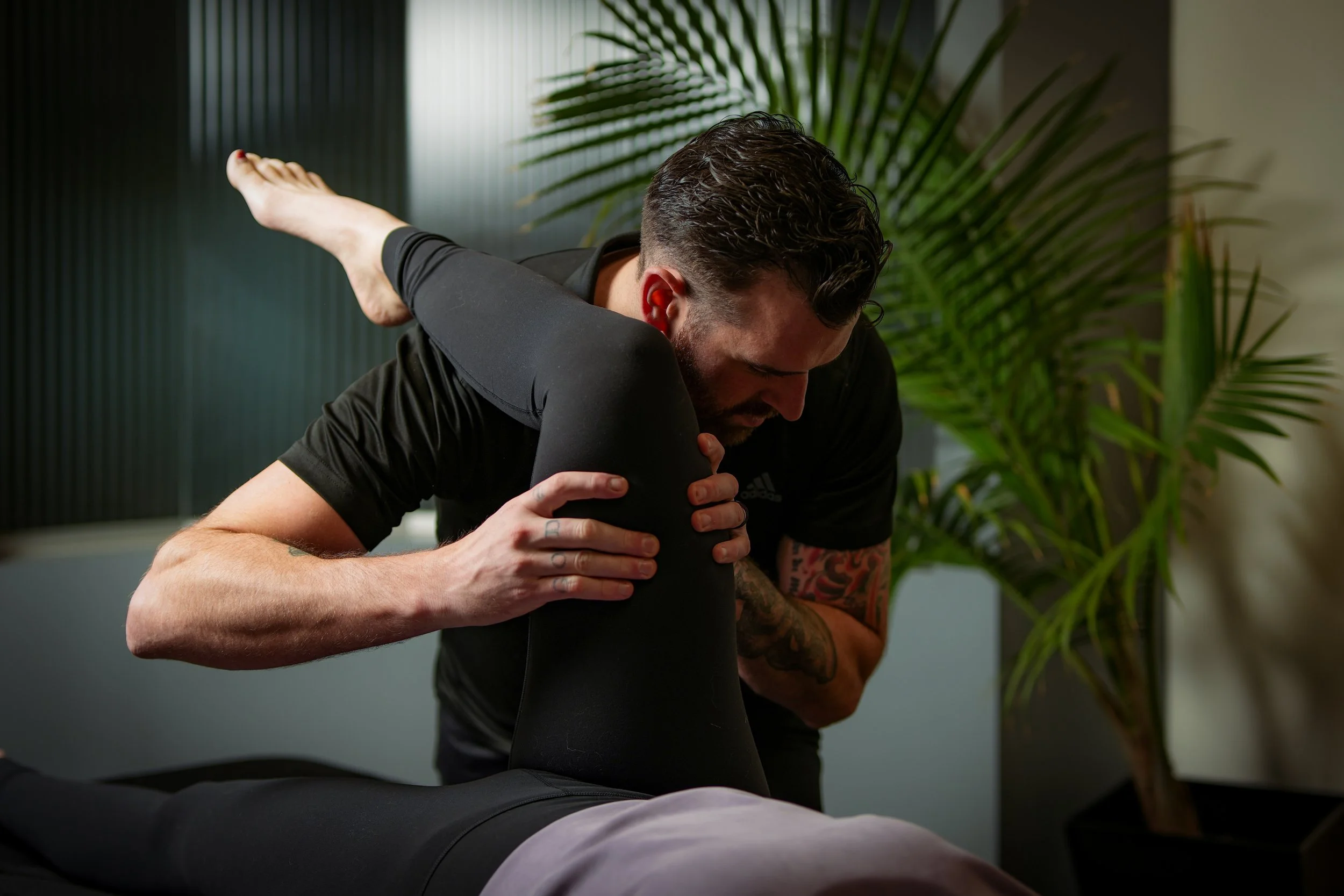 A man receiving physical therapy or massage on his shoulder from a therapist in an indoor setting with plants in the background.