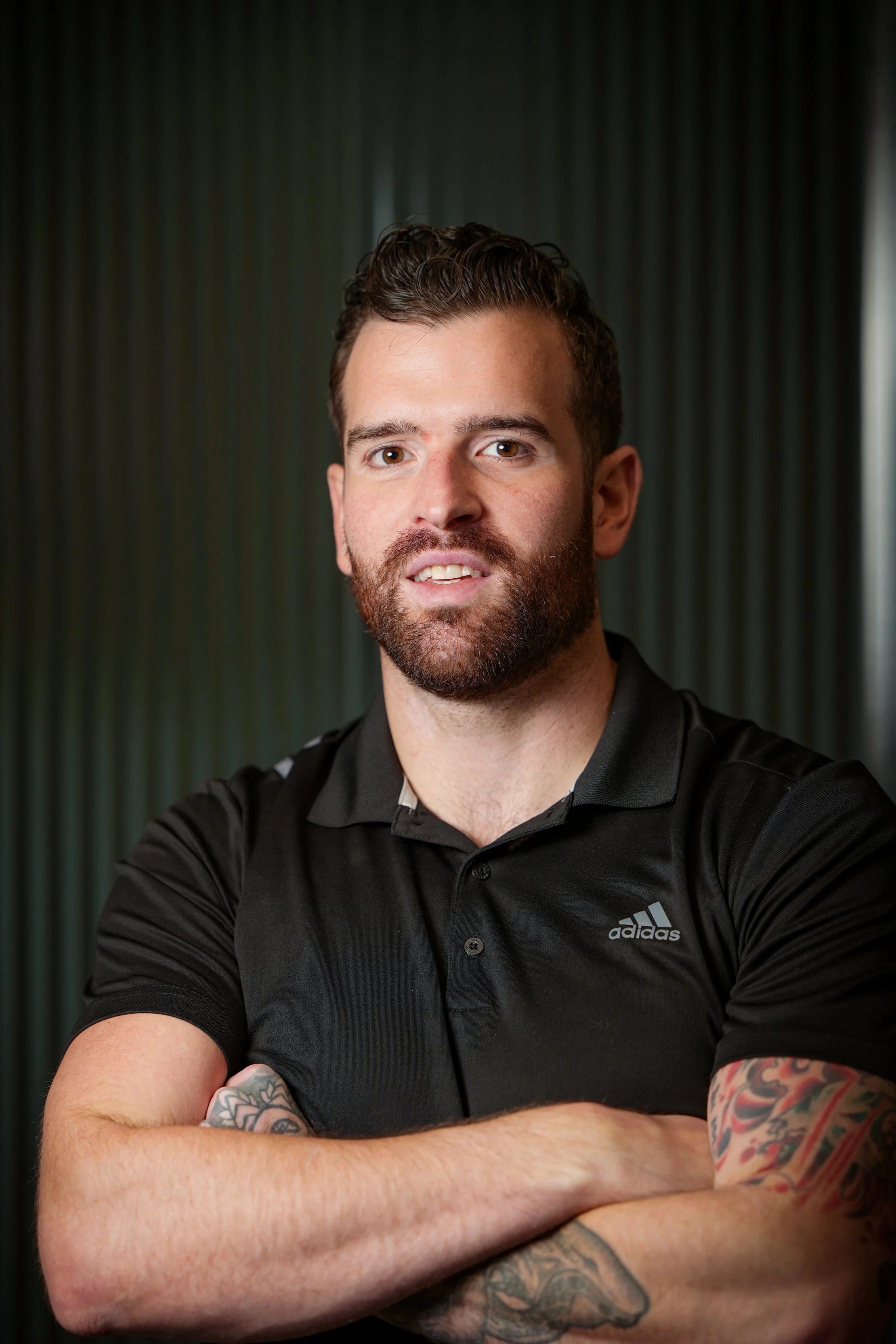 A man with tattoos on his arms, a beard, and mustache wearing a black Adidas polo shirt with arms crossed in front of a dark background.