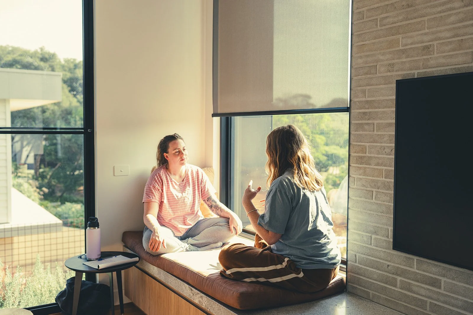 Two women sitting on a window seat, engaged in conversation, with large windows and green trees outside sunlight behind them.