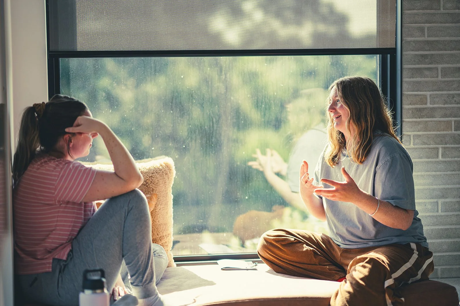 Two women sitting near a large window, engaged in a lively conversation. One woman is laughing, gesturing with her hands, while the other woman listens, covering her face with her hand. Sunlight filters through the window illuminating their discussion.