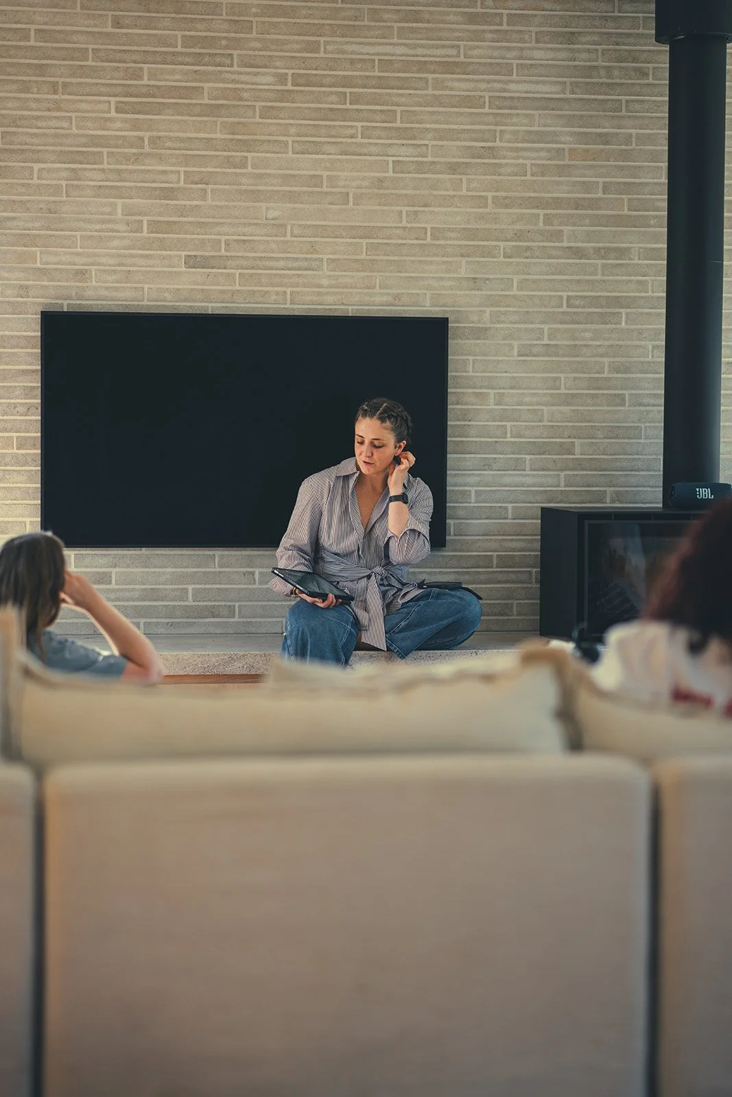 A woman sitting cross-legged on a low platform or shelf, holding a tablet and speaking to a group in a cozy living room with a brick wall, a large black screen, and a TV or speaker.