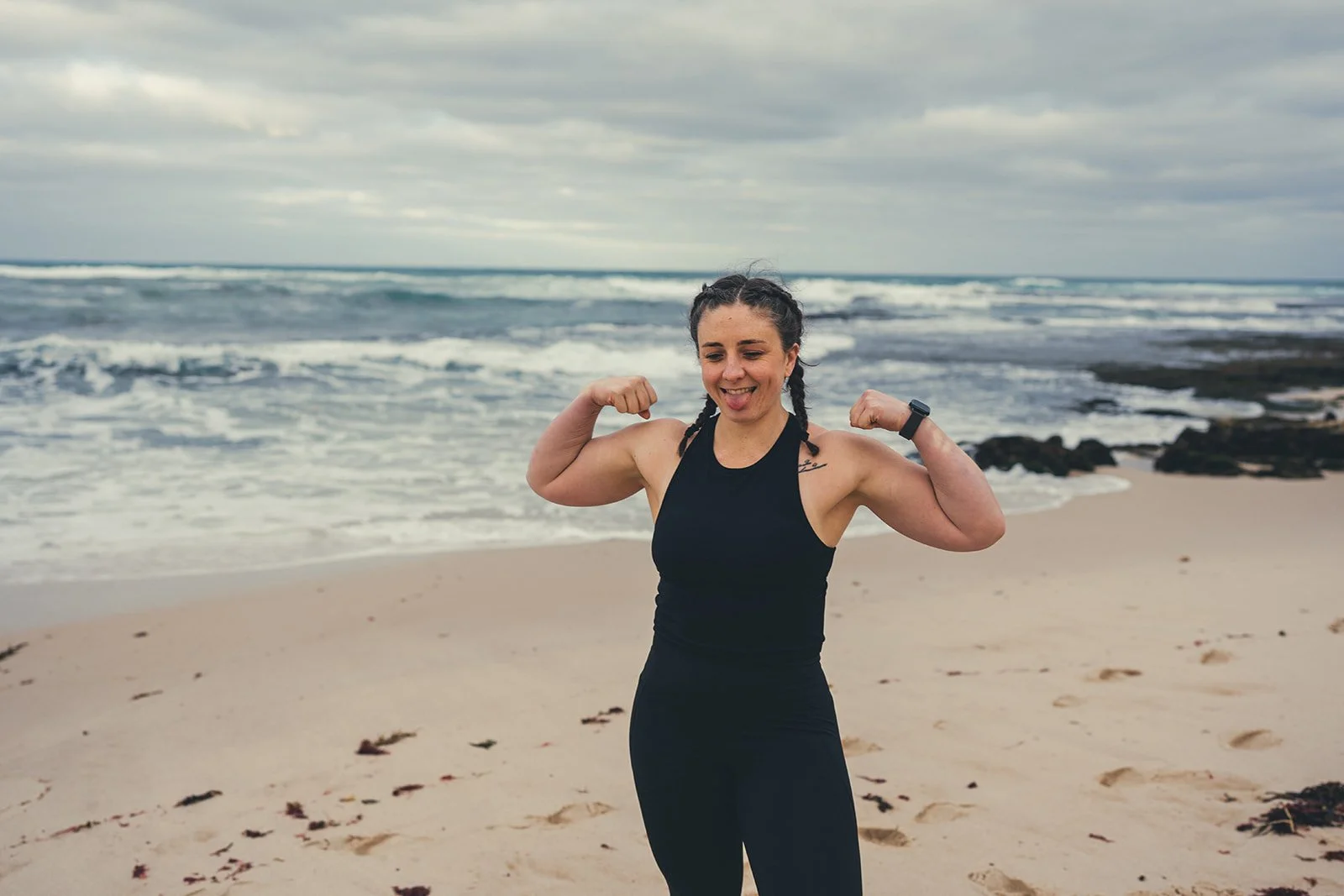 A woman with braided hair, wearing a black sleeveless athletic top and leggings, is flexing her biceps and smiling on a sandy beach with waves and a cloudy sky in the background.