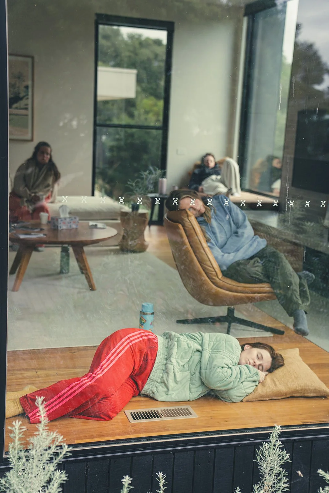 People resting inside a house, viewed through a window. A woman is sleeping on the wooden floor with a pillow, dressed in a light green jacket and red pants. Inside, others are seated or lying on chairs and couches near large windows showing greenery outside.