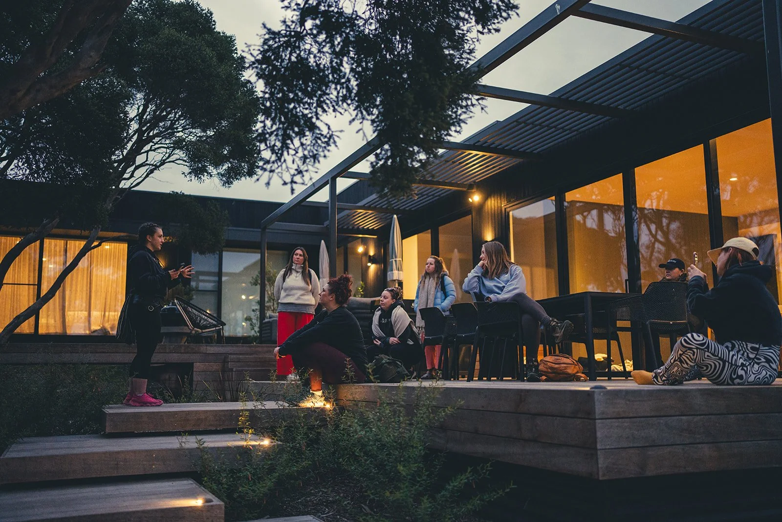 Group of people gathered outside a modern house with large glass windows during evening, with a woman speaking to the audience.