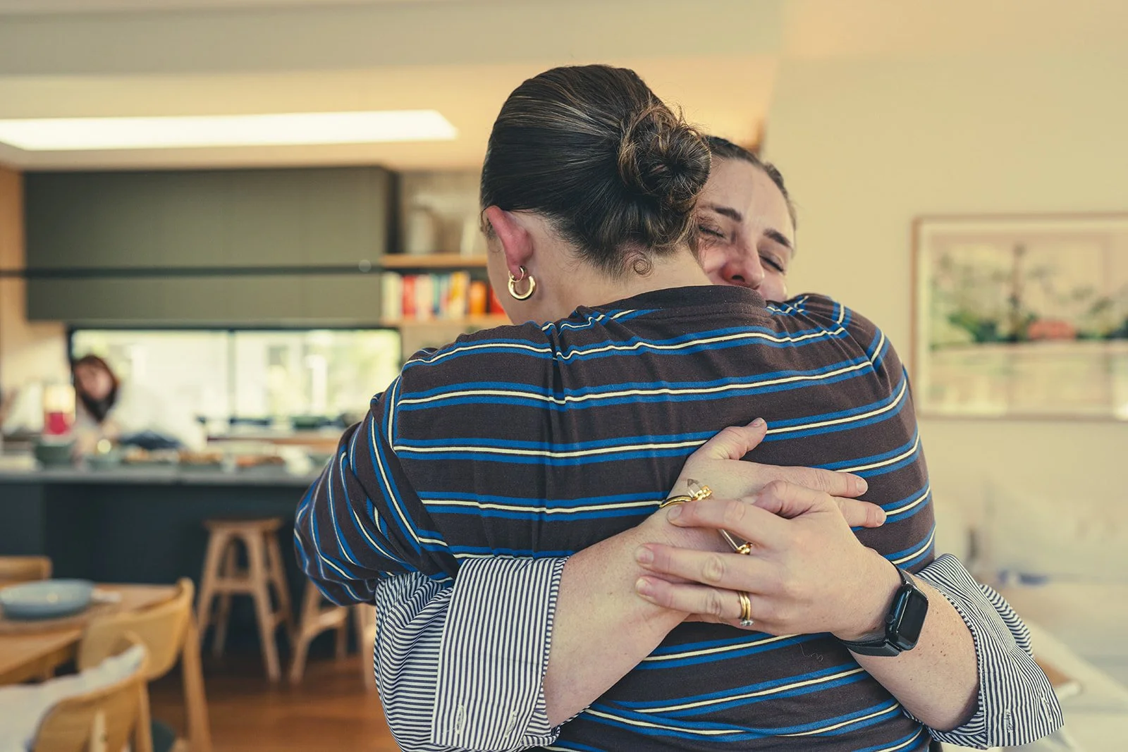 Two women hugging each other warmly in a cozy, well-lit room. One woman is wearing a striped shirt and gold jewelry, while the other woman is wearing a long-sleeve shirt and a watch. In the background, a person is working in a kitchen with shelves and books visible.