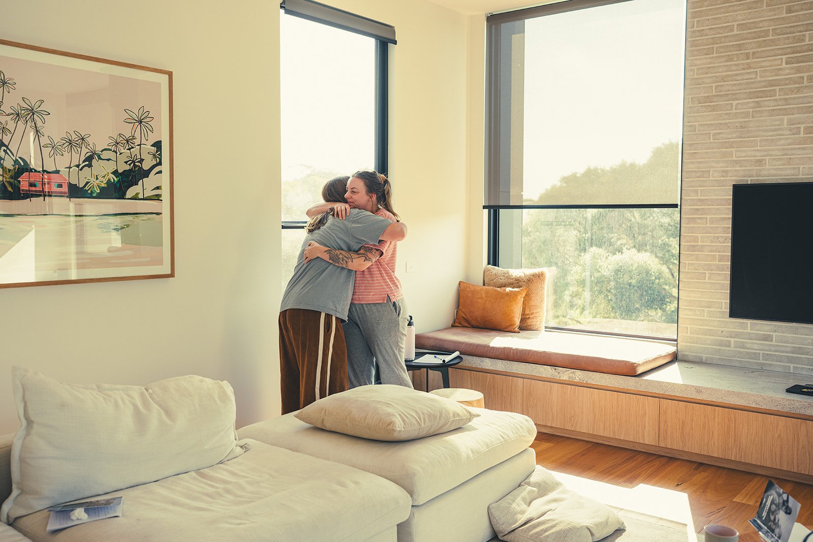 Two women hugging in a bright living room with large windows, a couch, and wall art.