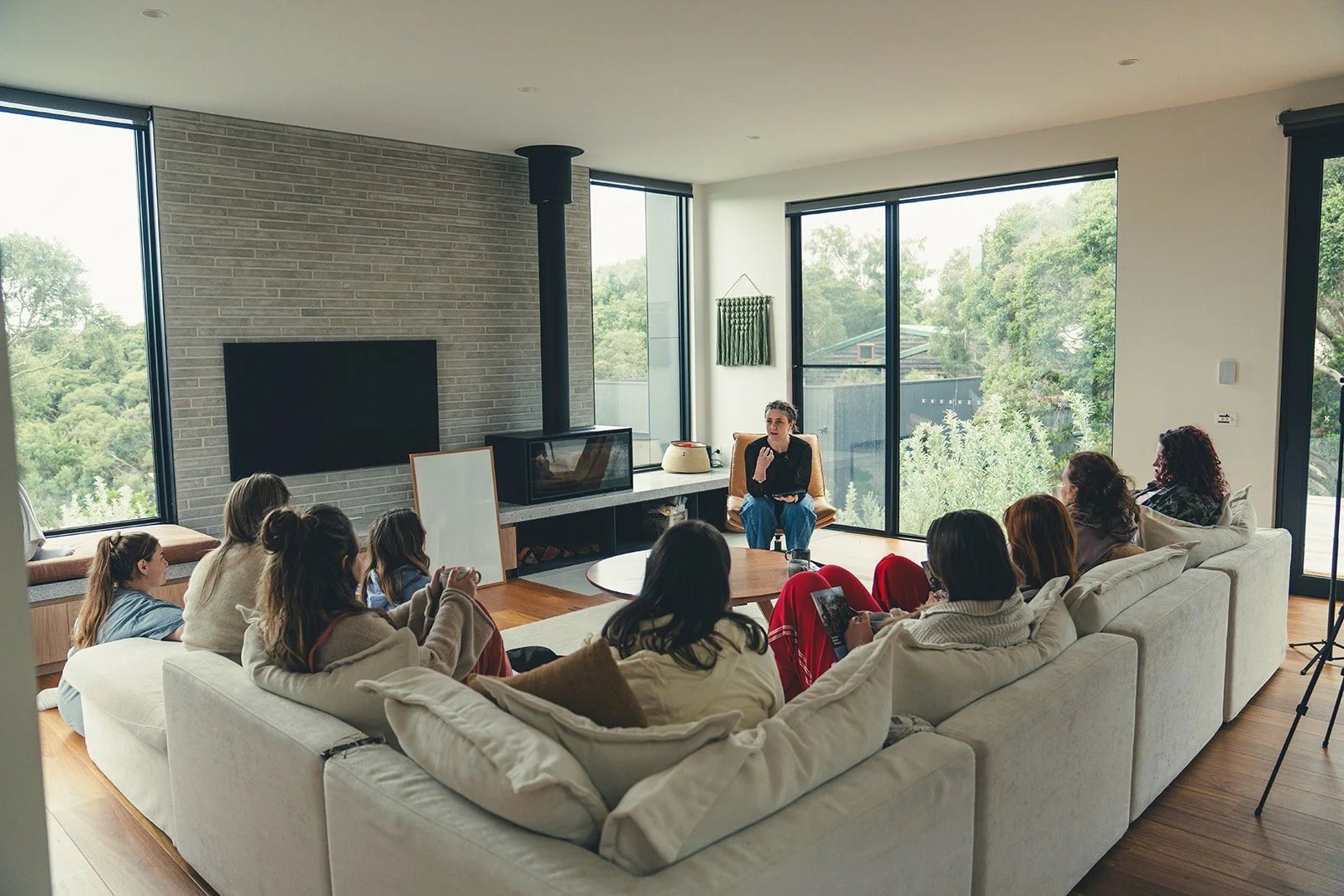 A woman leading a discussion or storytelling session for a group of children and adults sitting on a large white sectional sofa in a bright living room with large windows and a view of trees outside.