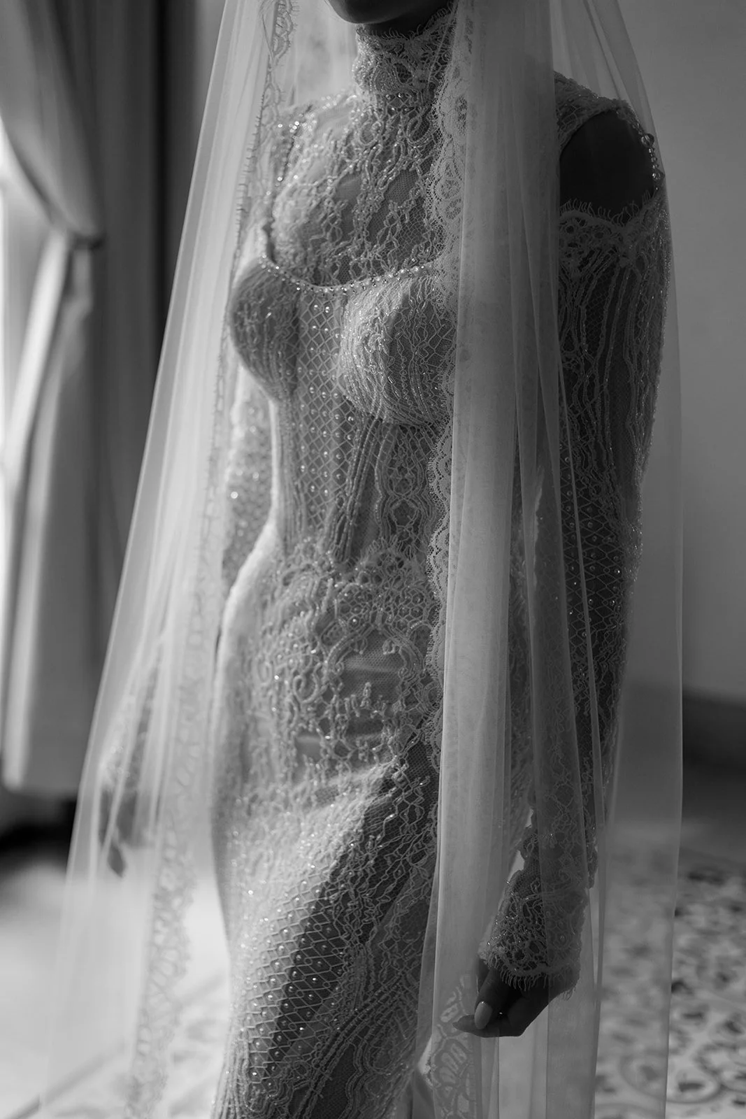 Black and white photo of a woman in a lace wedding dress, partially covered by a sheer veil, standing near a window.
