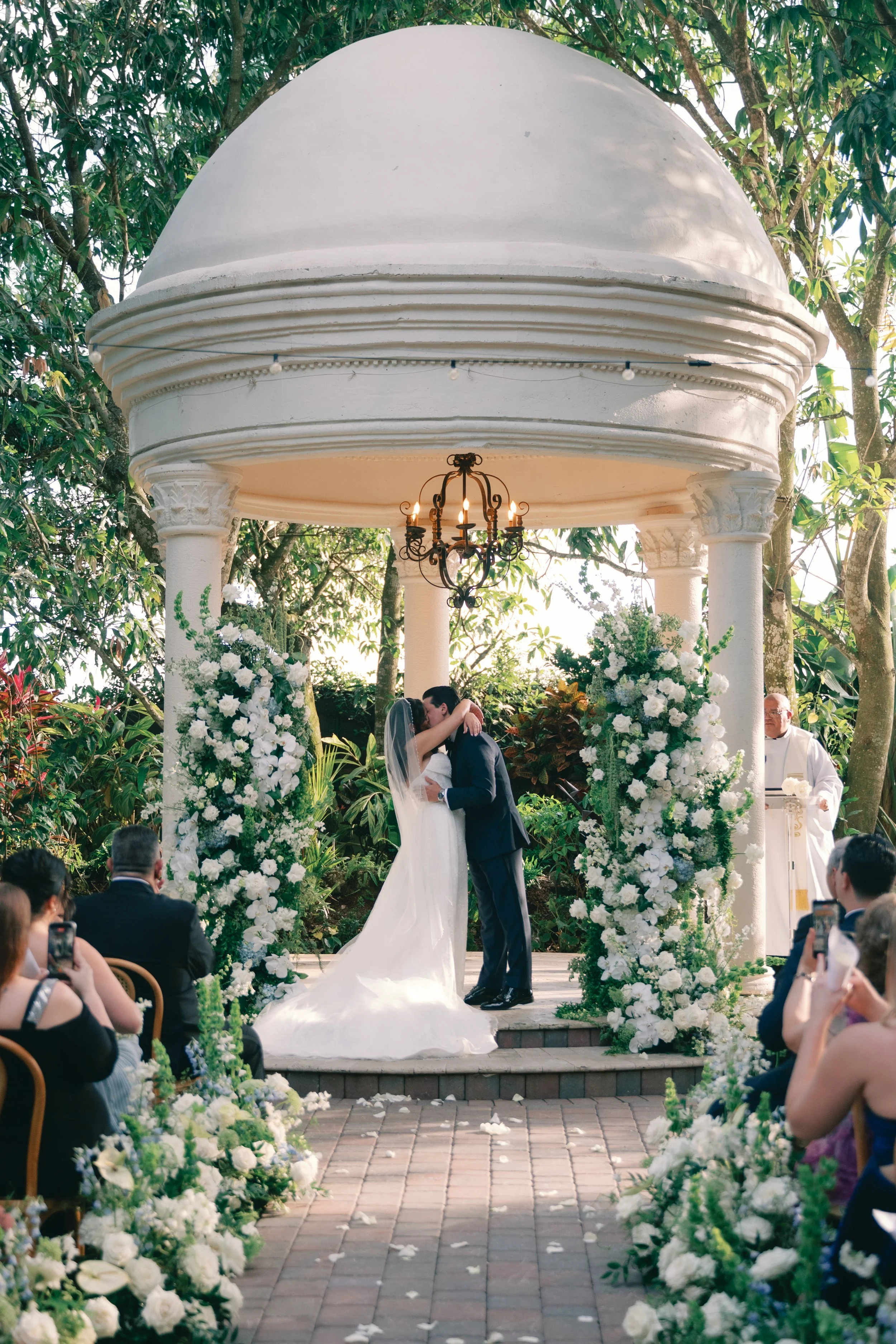 A couple sharing a kiss during their wedding ceremony under a white gazebo decorated with white flowers, surrounded by green trees and seated guests, some taking photos.