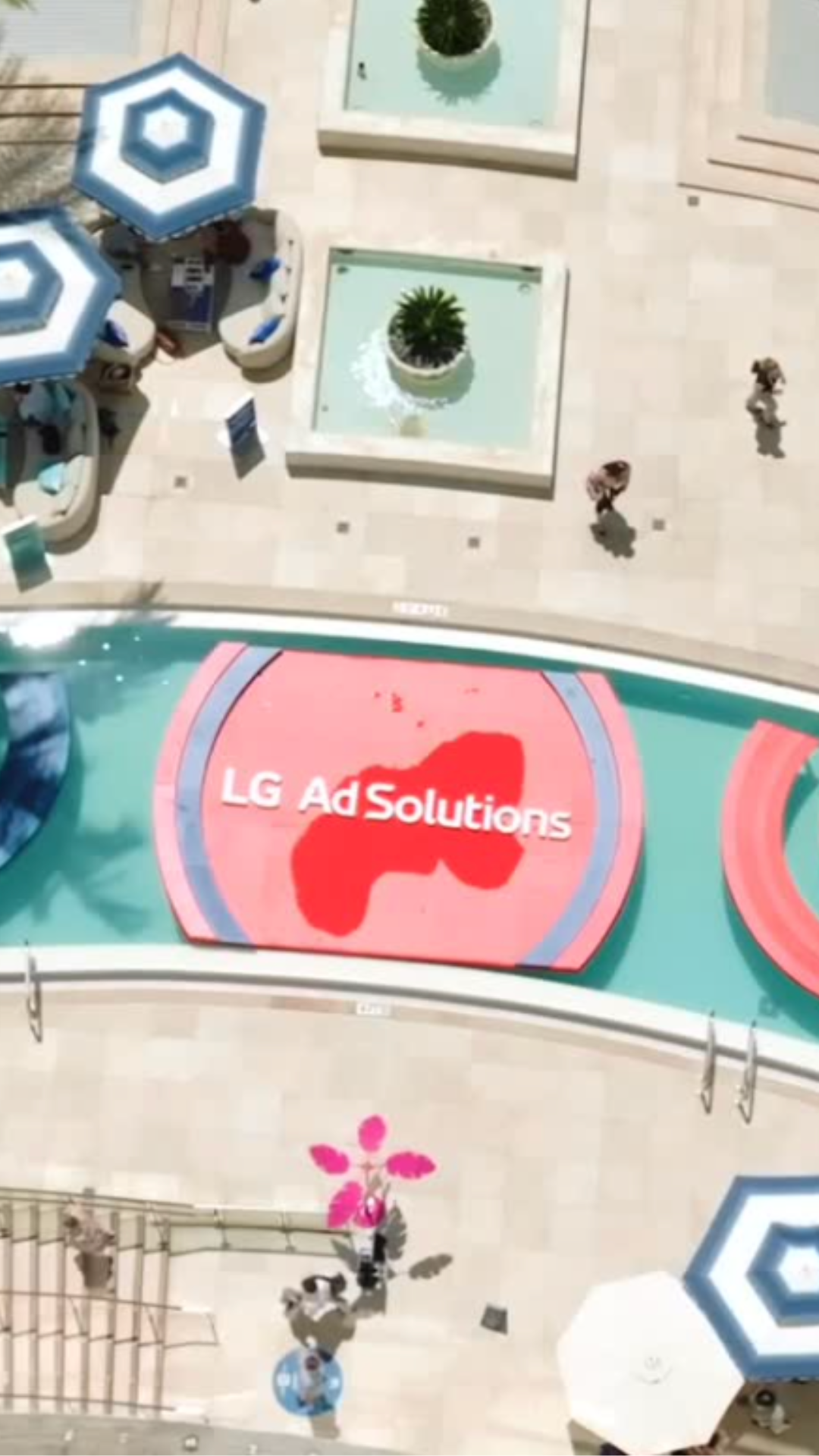 Aerial view of a pool area with a large red sign that reads 'LG AdSolutions' and depicts the shape of a country, surrounded by people walking and white umbrellas.