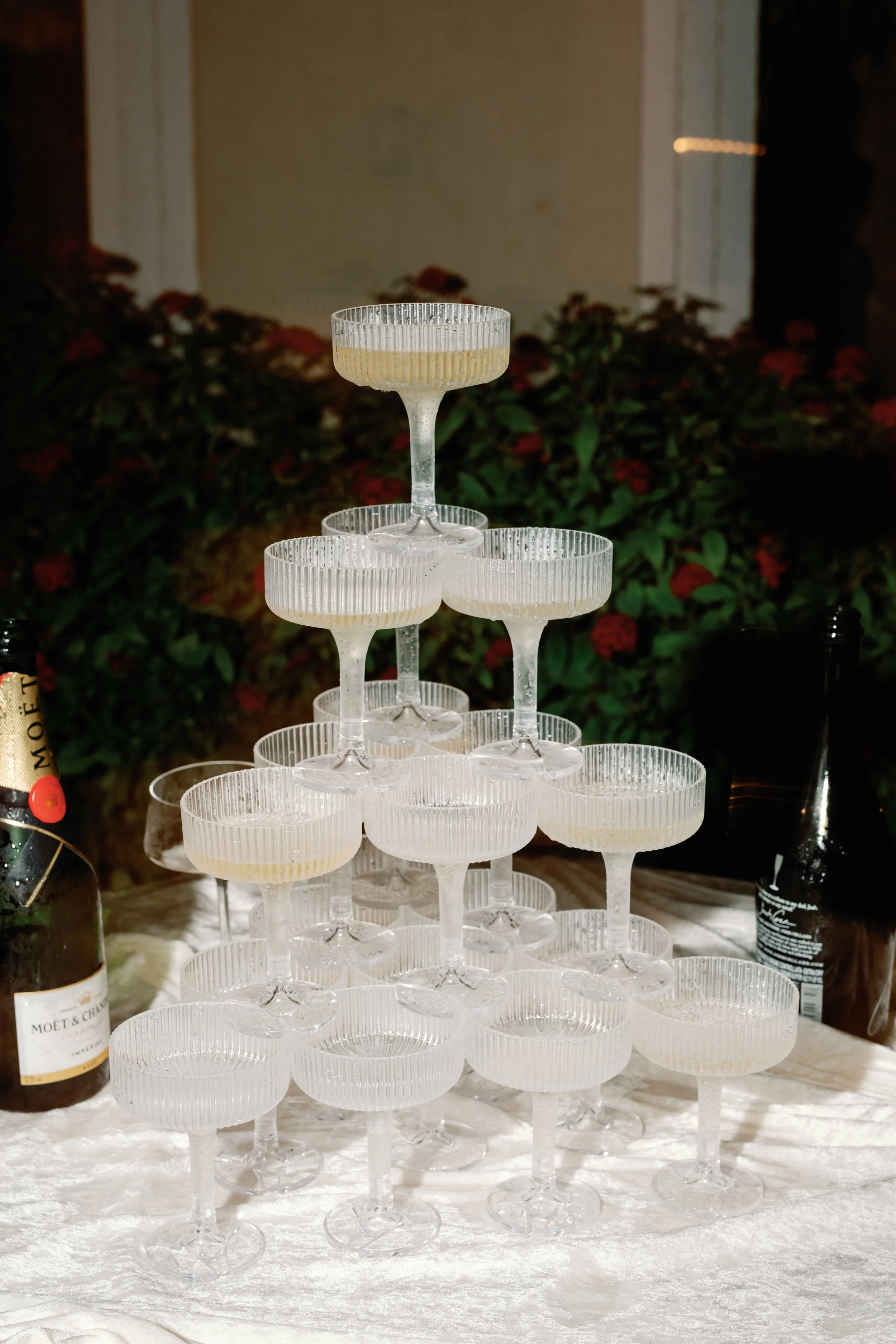 A pyramid of empty champagne glasses stacked on a white tablecloth, with bottles of champagne nearby, against a background of bushes with red flowers at night.