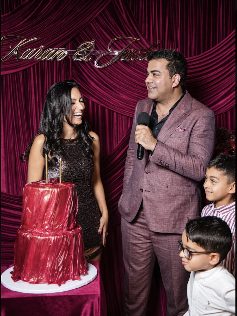 A woman and a man are celebrating at a party with a red cake, with a purple curtain backdrop that has gold cursive lettering.