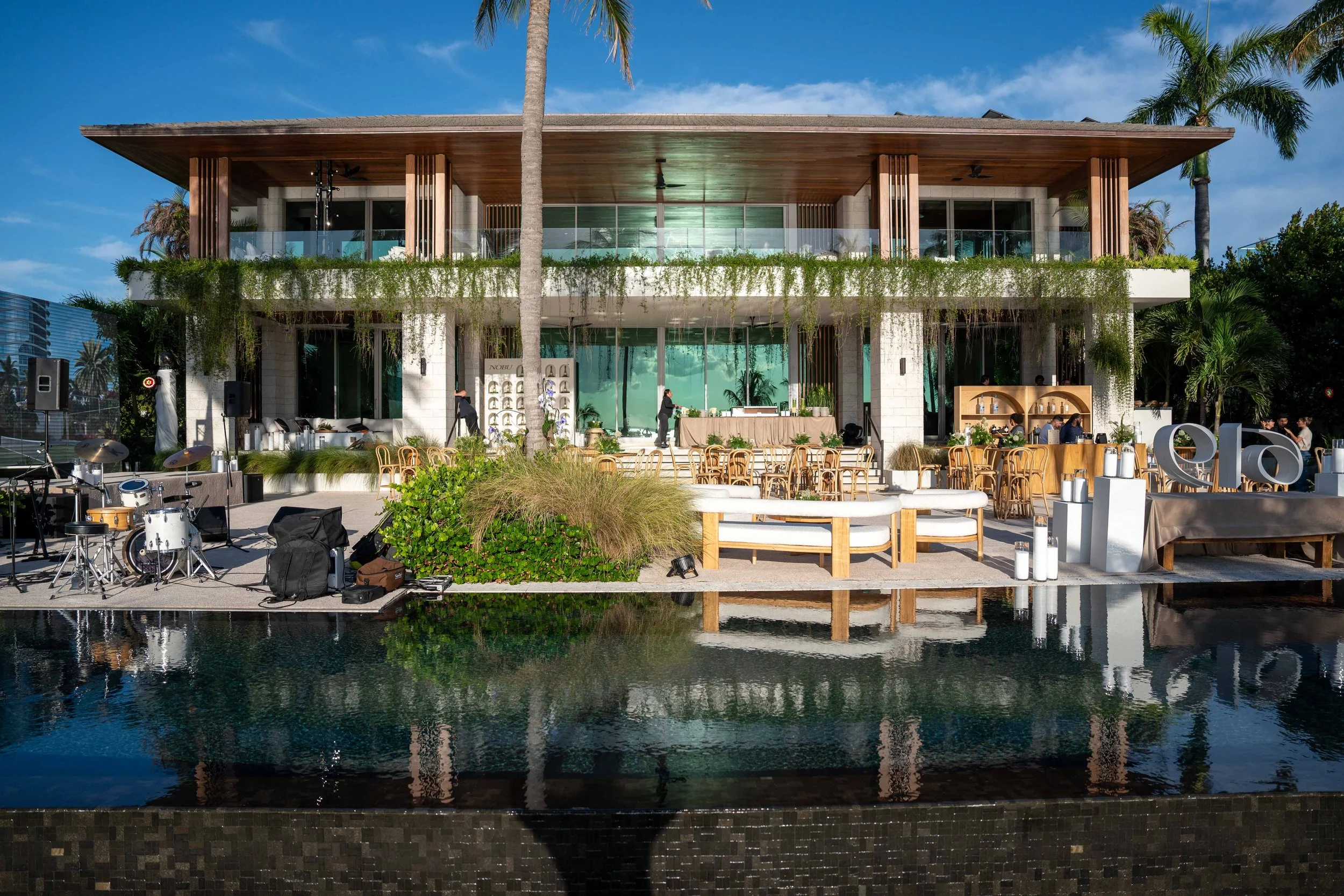 A modern two-story house with a large wooden overhang, glass balcony, and lush greenery on the balcony and around the house, located near a water body with a reflection of the house.