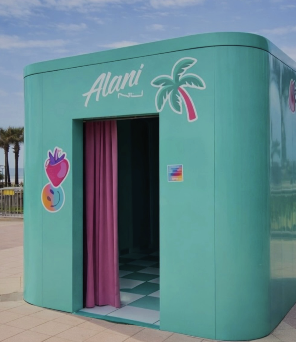 A teal-colored photo booth with a pink curtain, decorated with colorful tropical fruit and drink logos, and the word 'Alani' on top, located outdoors near palm trees and a sidewalk.