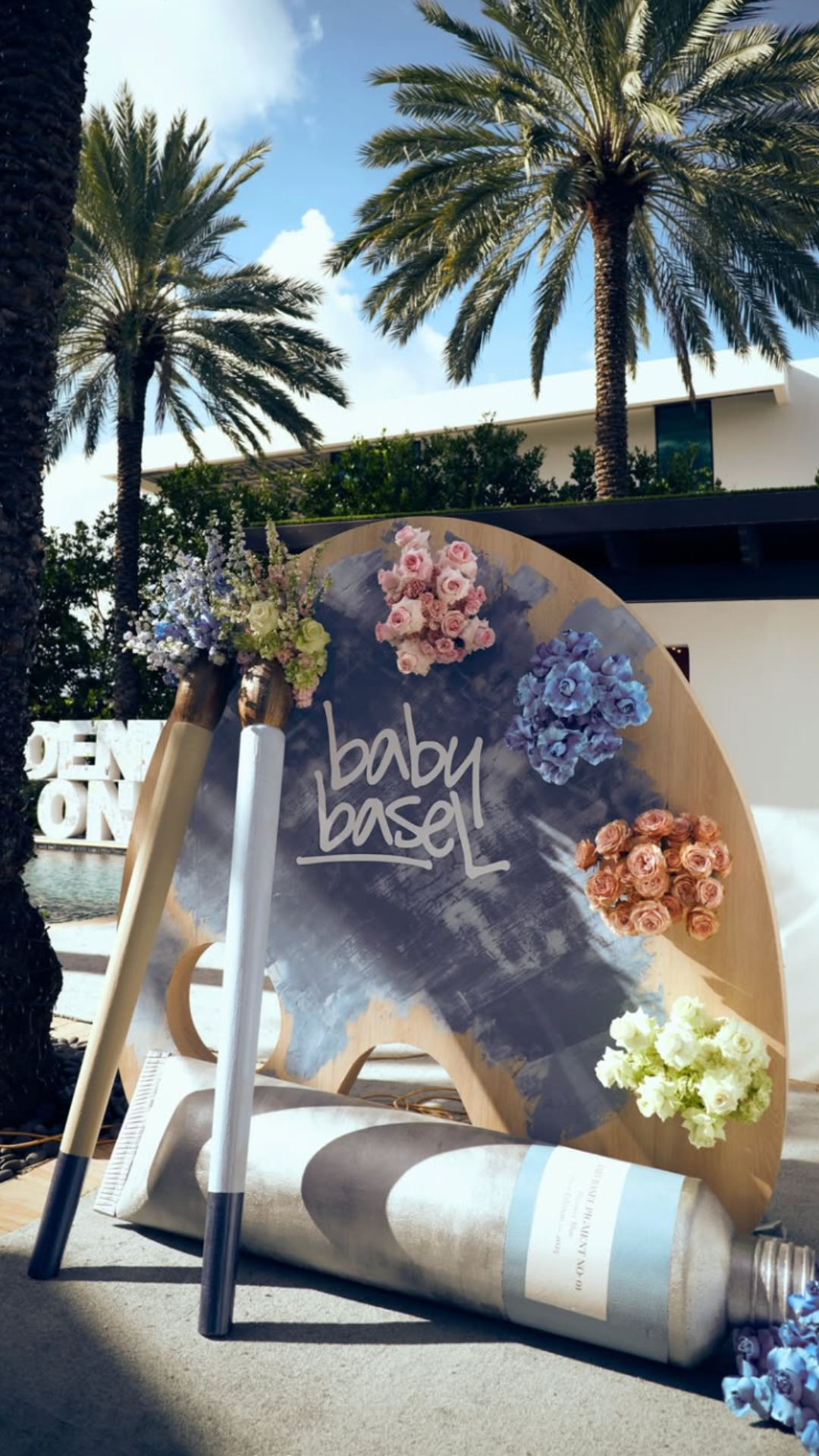 Outdoor scene with palm trees and a decorative sign reading 'baby basin,' decorated with pink, purple, peach, and white flowers, and a large illustration of a baby bottle.