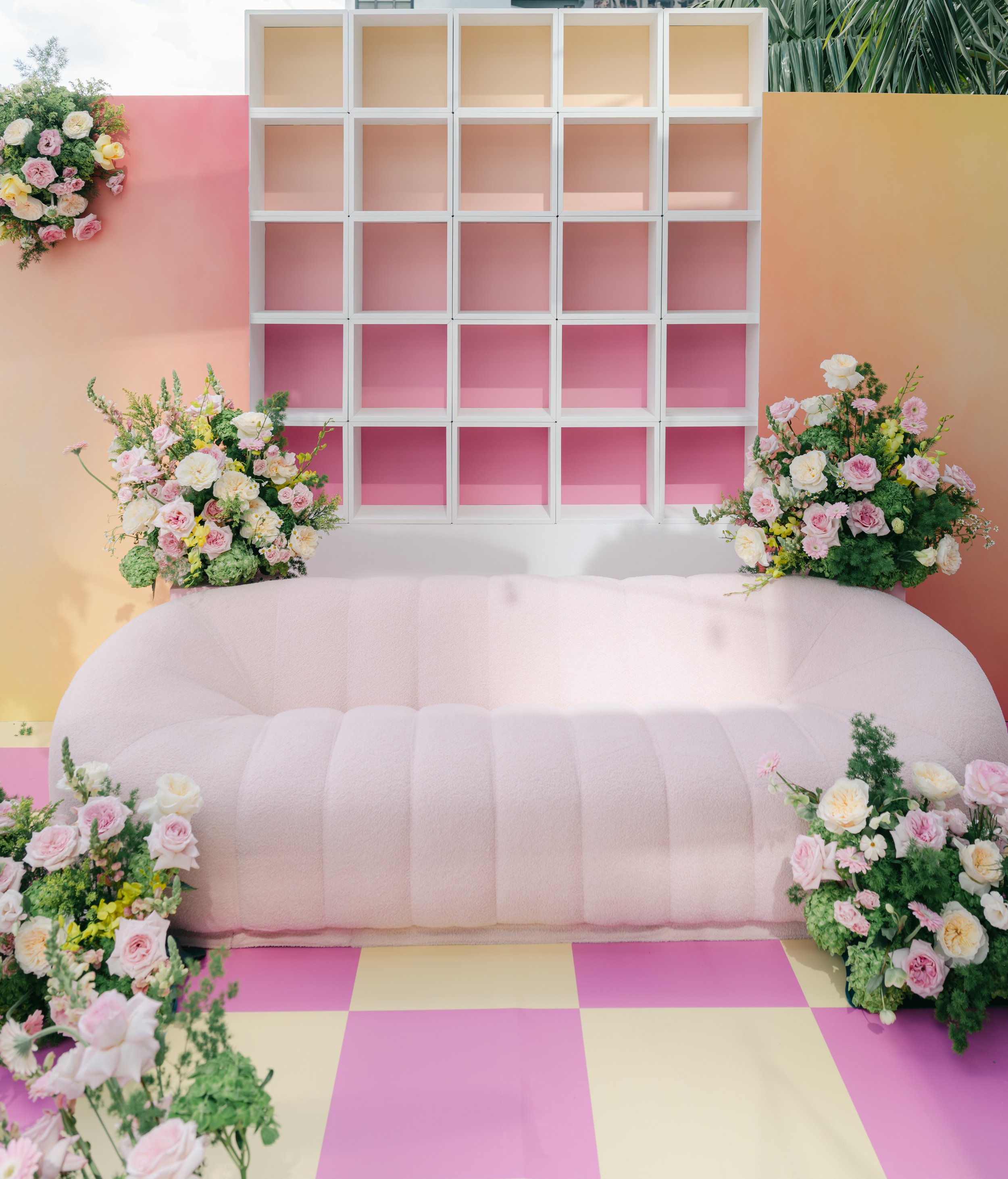 Pink and yellow checkered floor, white cushioned seating, pink and beige wall with white grid shelving, and floral arrangements with pink, white, and yellow flowers.