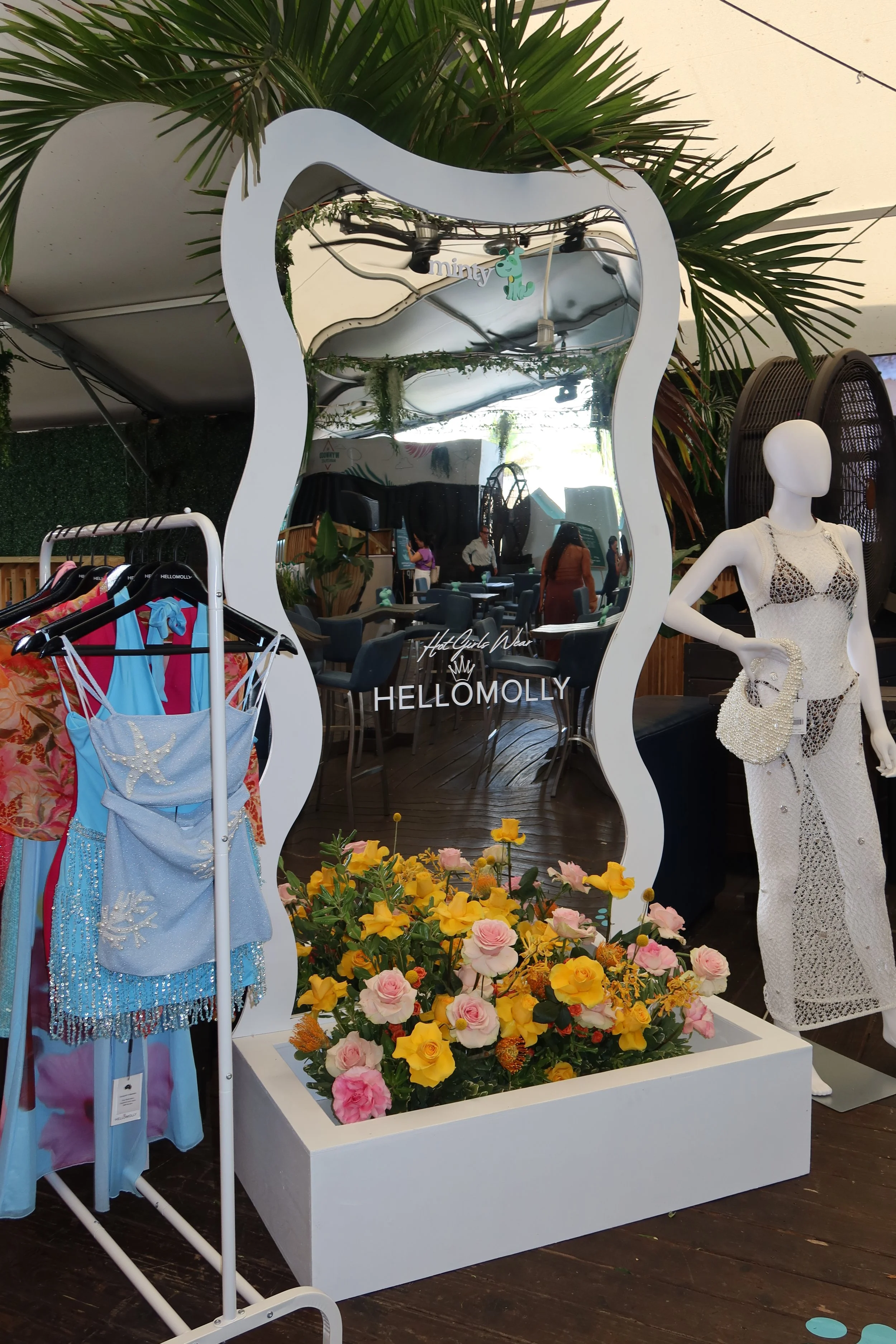 A store display with a white, irregularly shaped mirror, a mannequin wearing a white lace dress, a rack of colorful clothes, and a large flower arrangement with yellow and pink flowers in front of the mirror. There are green plants and seating inside