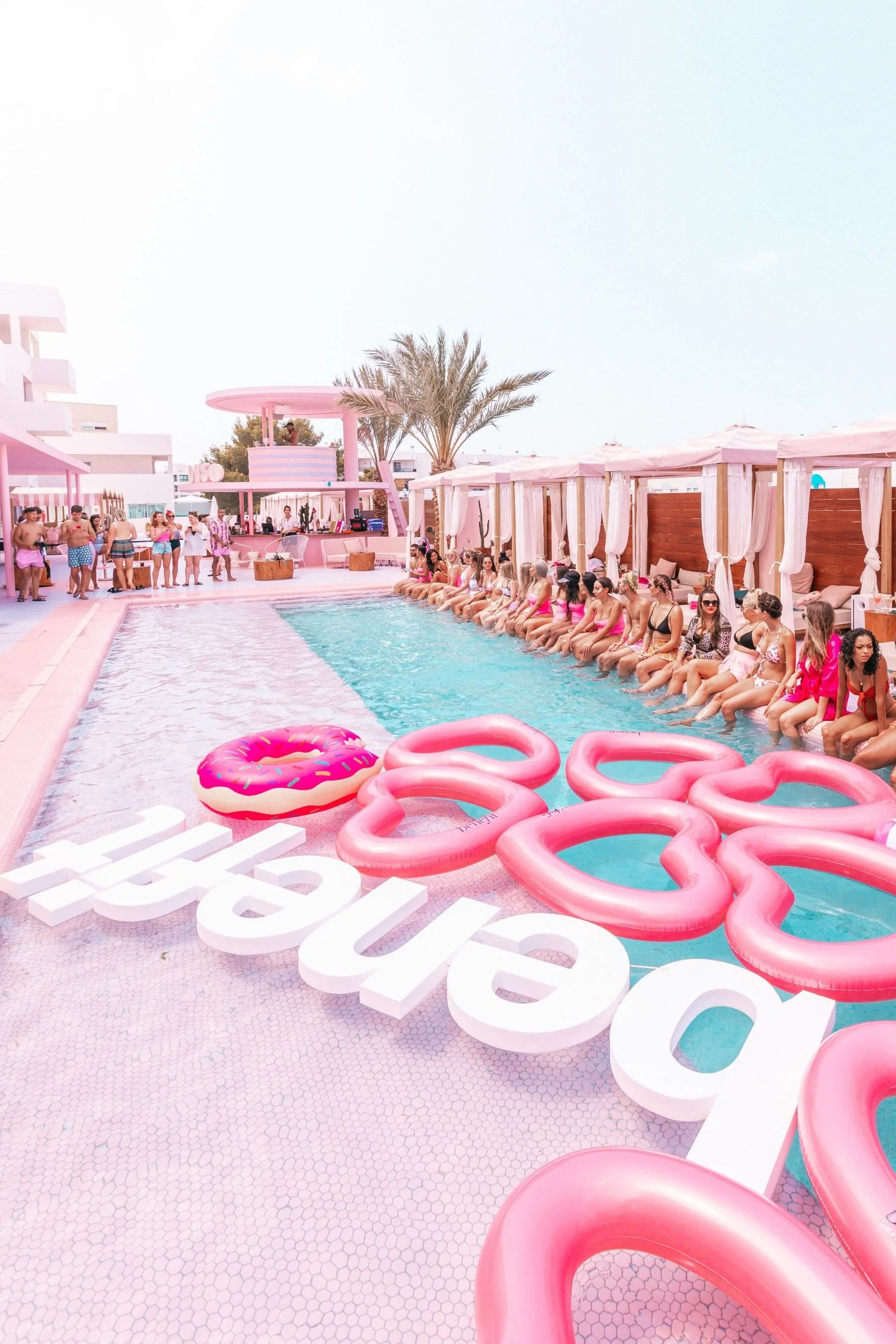 People at a poolside bar with pink décor, including pool floats shaped like pink donuts and pink flowers, with seating areas featuring white curtains and umbrellas.