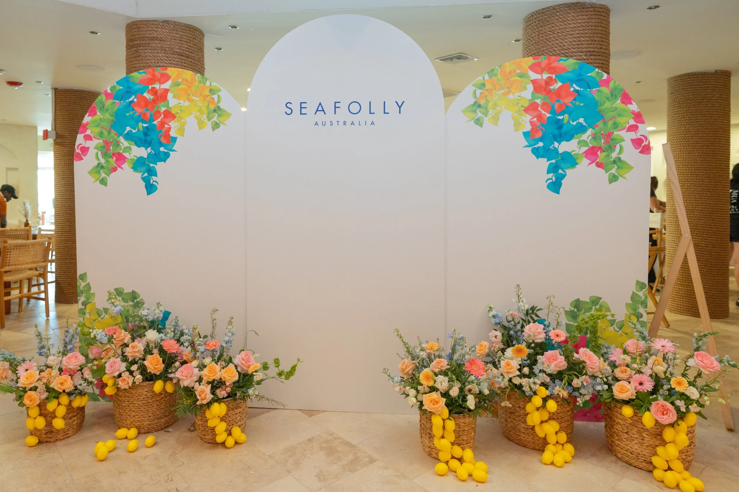 Decorative floral display with baskets of pink, yellow, and pastel flowers, and yellow lemons, surrounding a white backdrop with the sign 'SeaFolly Australia' at a retail or event location.
