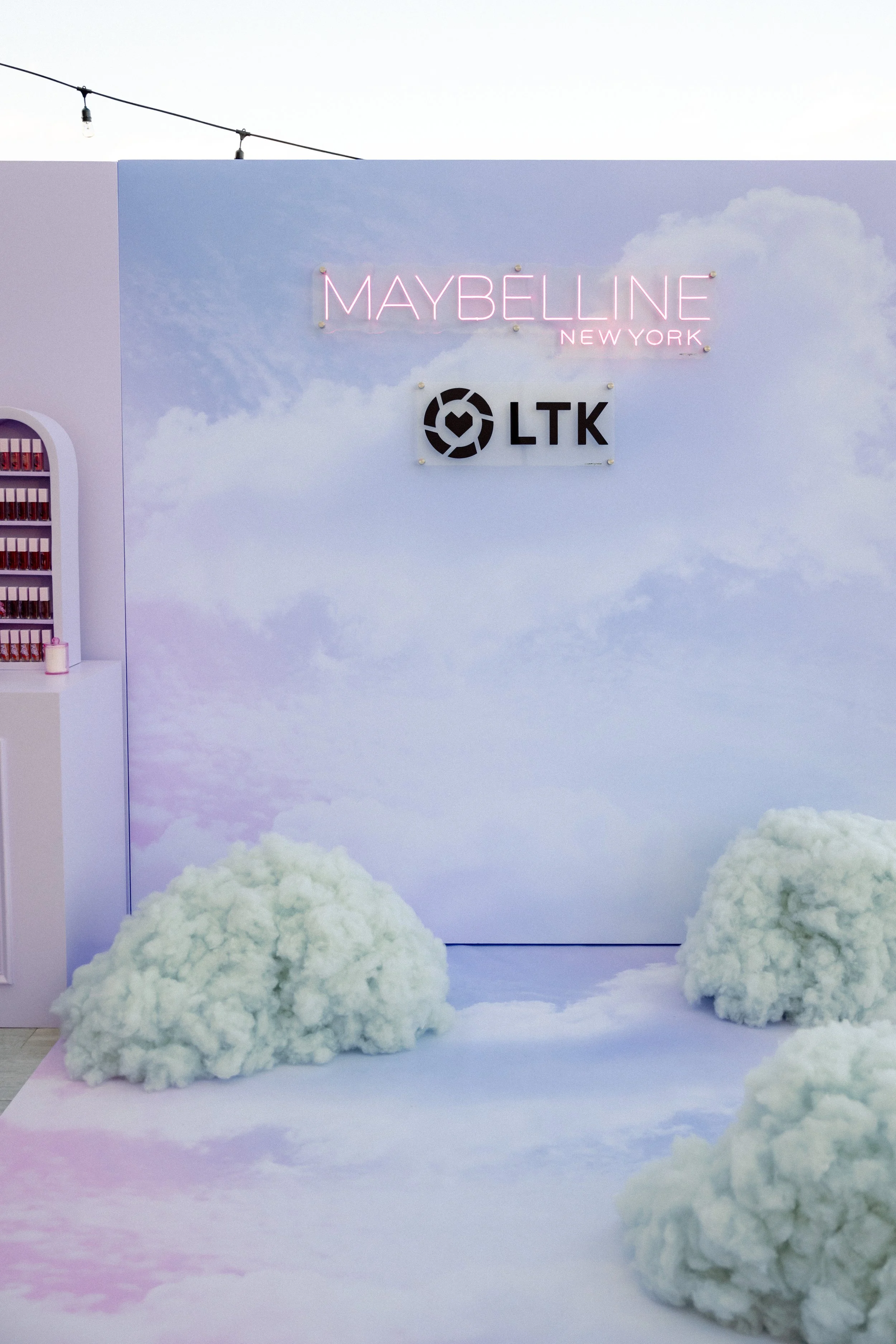 A display with a pastel sky background, plush cloud props on the floor, a table on the left with makeup products, a pink neon sign saying 'Maybelline New York', and a logo with the letters 'LTK' and a heart icon.