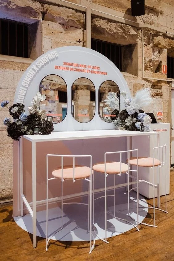 Beauty display setup with three white bar stools in front of a pink counter, decorated with flower arrangements and a mirror with a white frame, on a wooden floor against a stone wall.