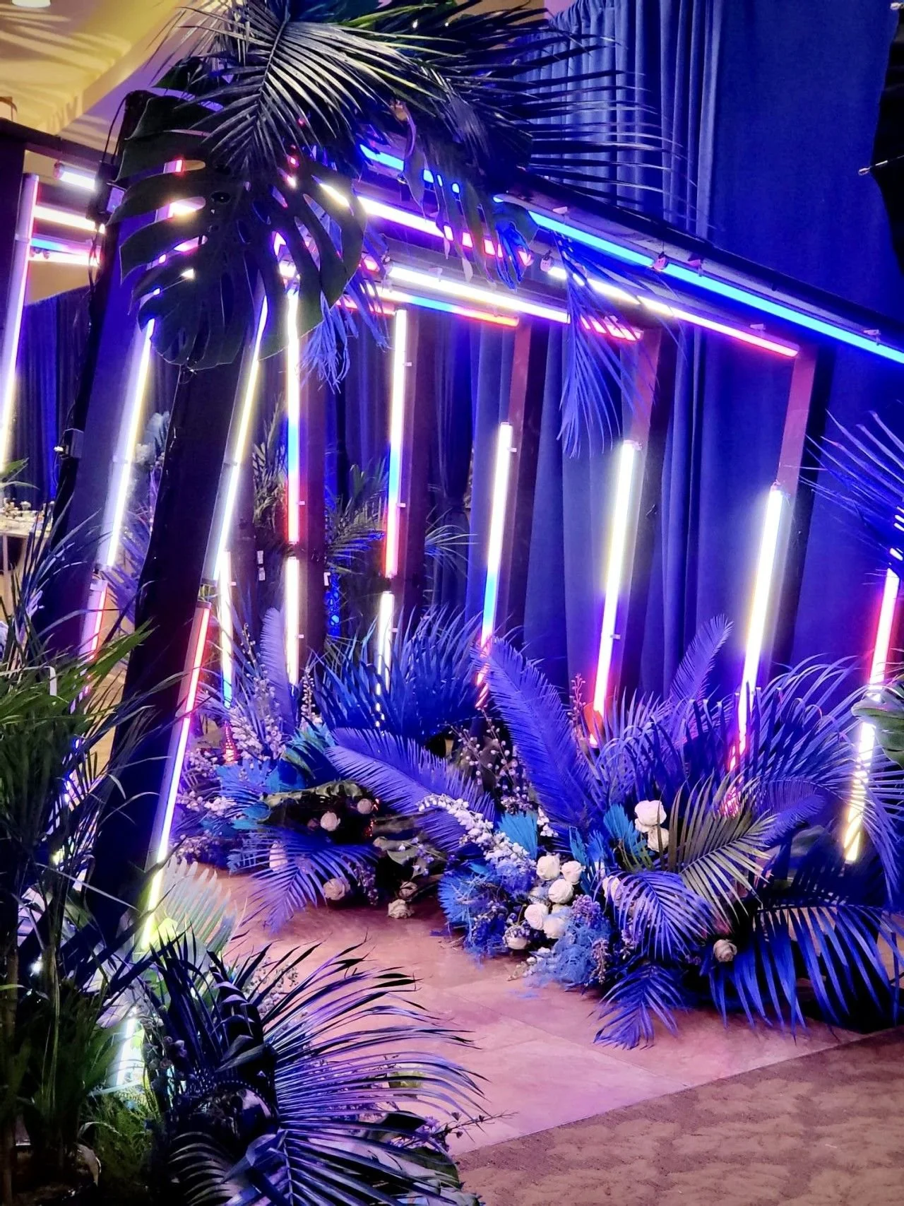 A decorative scene with tropical plants illuminated by neon lights in pink, purple, and blue, set against dark curtains.