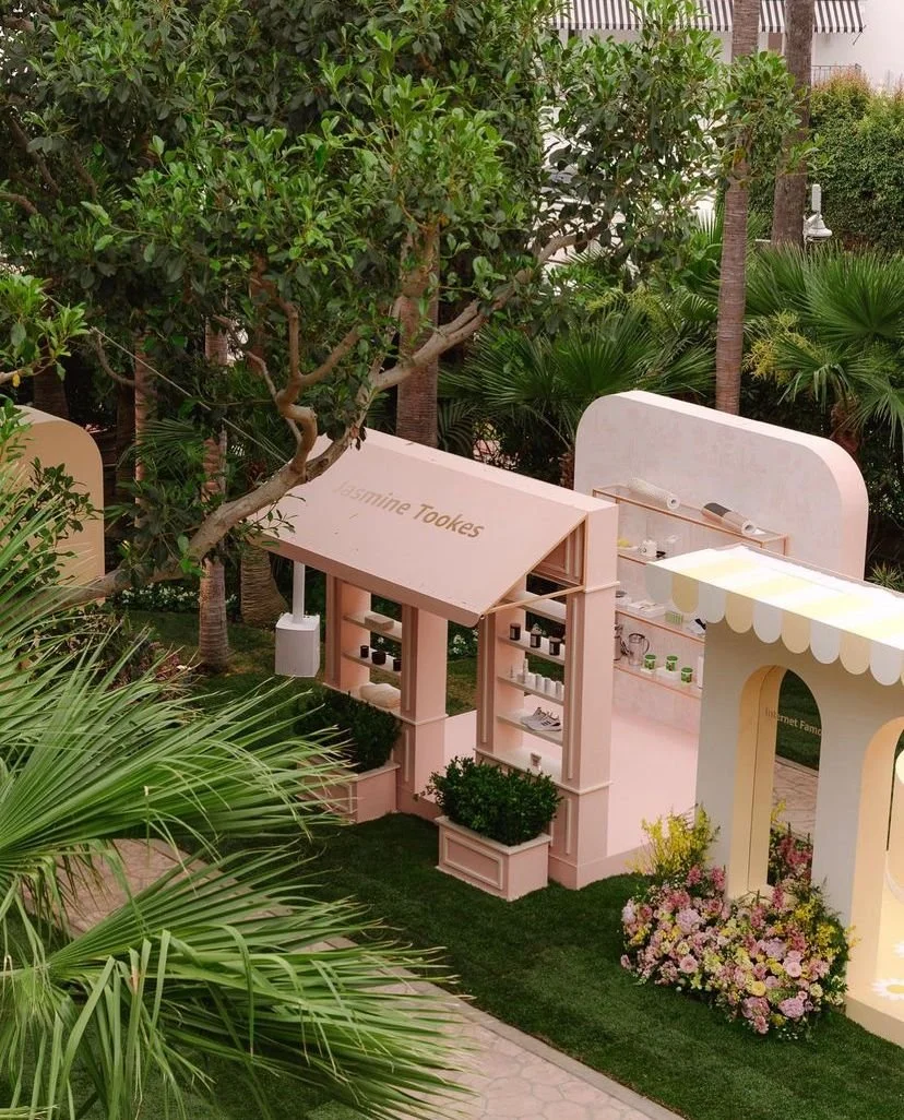 Miniature outdoor market setup with pink and beige structures, shelves, and flower arrangements among lush greenery and trees.
