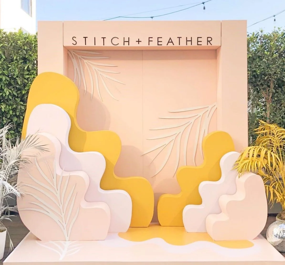 Decorative backdrop with beige and yellow abstract shapes, leafy patterns, and the logo 'STITCH + FEATHER' at the top, surrounded by potted plants.
