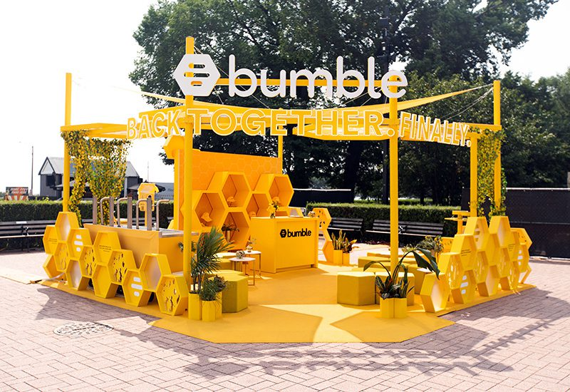 Outdoor promotional setup for Bumble with yellow decorative furniture and signs, featuring the Bumble logo and slogans like 'Back Together Finally,' surrounded by trees and a paved area.