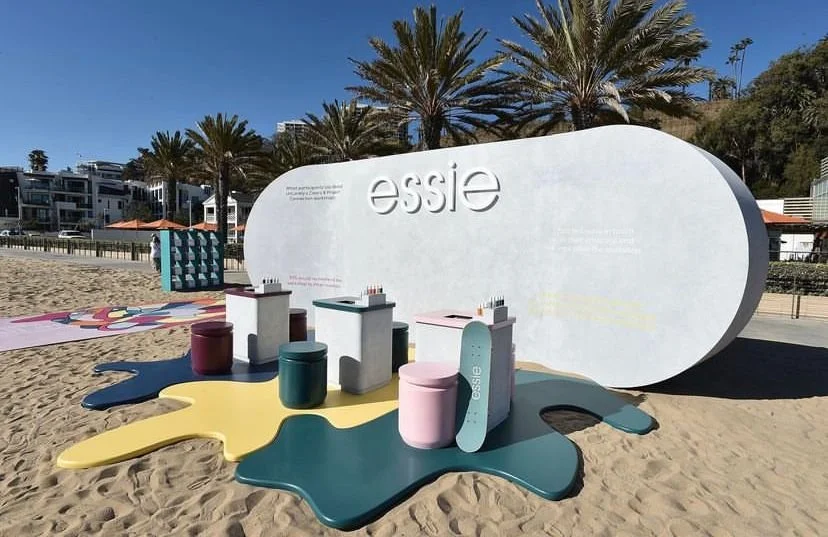 An outdoor display featuring the brand 'Essie' with colorful furniture and supplies on a sandy beach, palm trees, and modern buildings in the background.