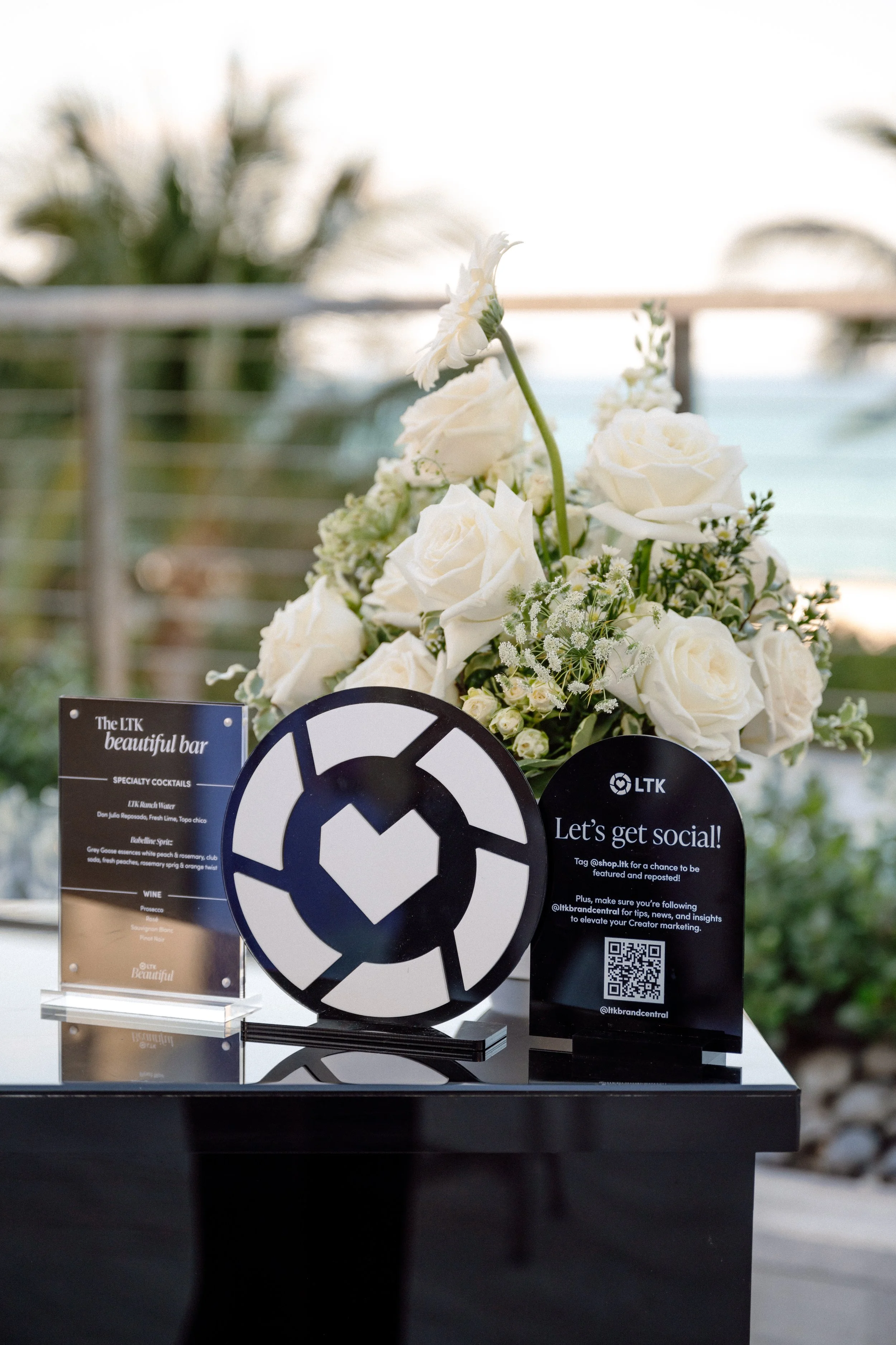 A bouquet of white roses and small white flowers on a table with signs promoting social media engagement and a QR code, set outdoors with a blurred background of trees and a railing.