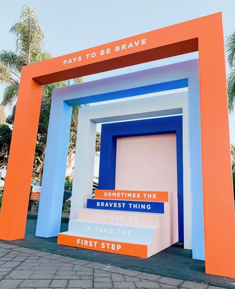 Colorful archways with motivational quote. Orange and blue frames with a staircase-like structure. The quote reads: 'PAYS TO BE BRAVE' at the top, and 'SOMETIMES THE BRAVEST THING YOU CAN DO IS TAKE THE FIRST STEP' on the steps.