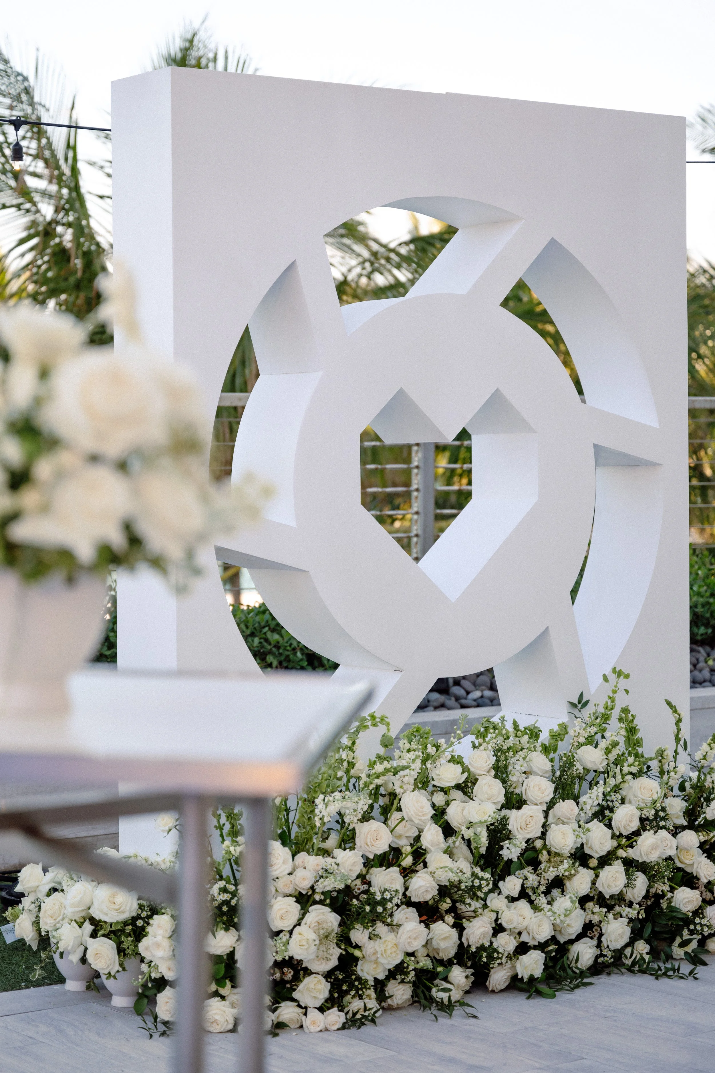 White floral arrangement with white roses and greenery at a wedding or event, with a large white geometric sculpture in the background and a blurred white banner or structure in the foreground.
