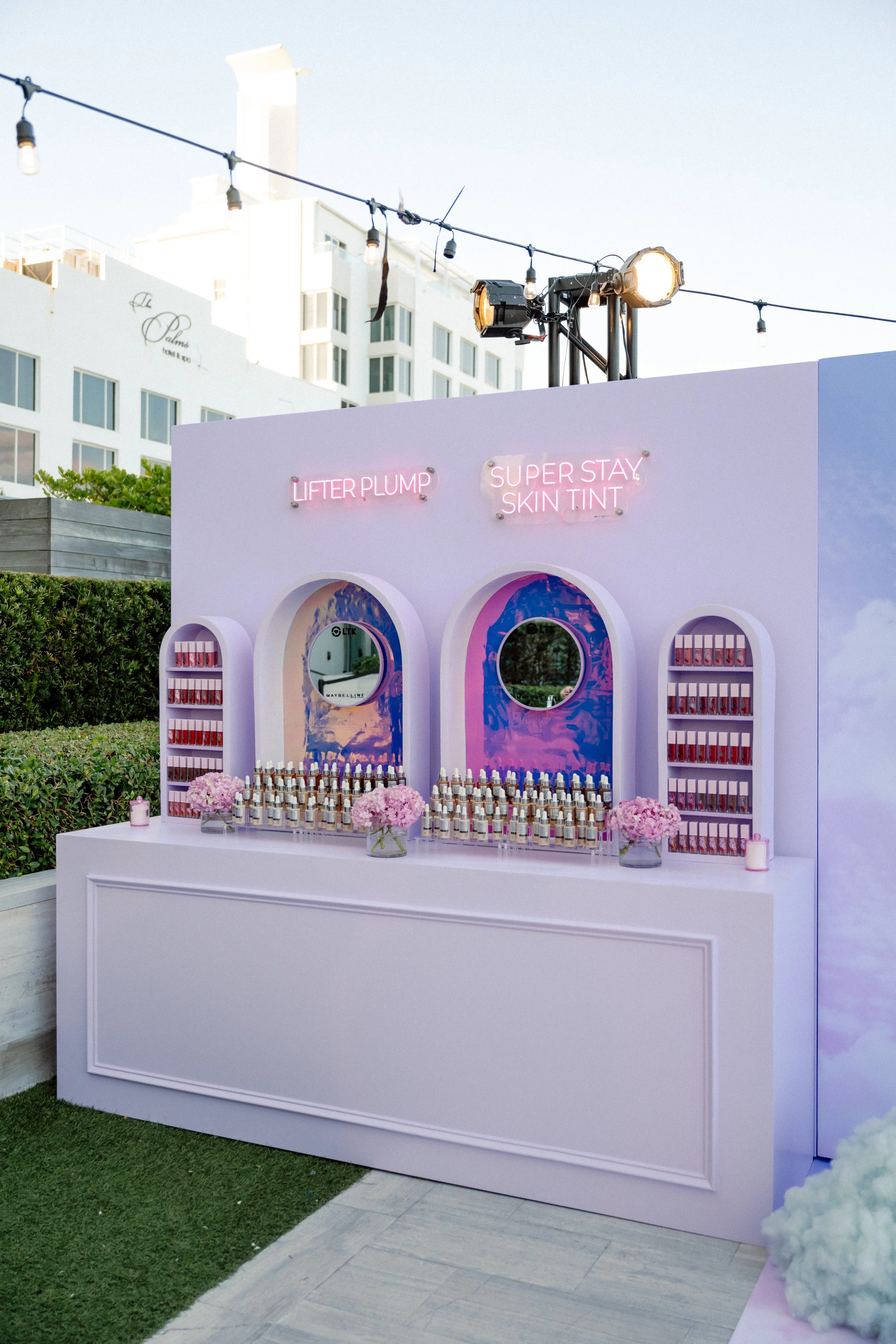 A pastel pink beauty product display with neon signs that say "Lifter Plump" and "Super Stay Skin Tint". The display includes bottles of nail polish, mirrors, flower arrangements, and is set outdoors with a building and string lights in the backgroun