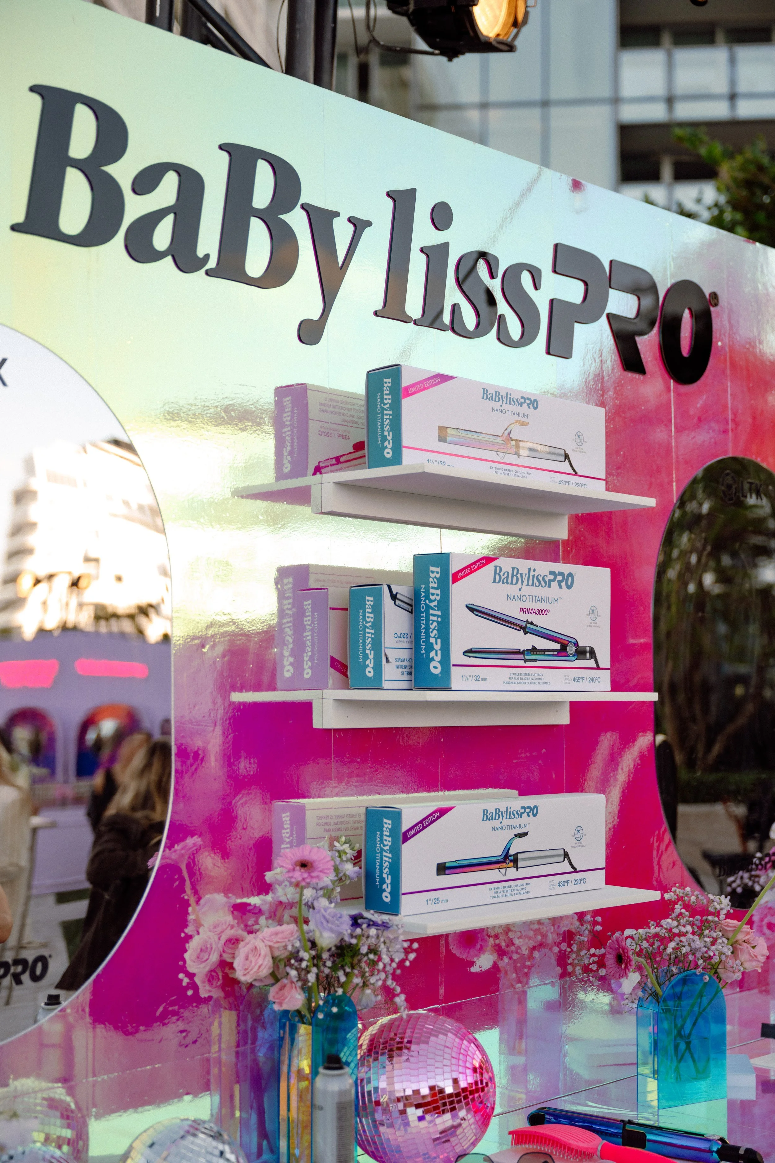 A colorful display of hair styling tools and accessories, including boxed BabylissPRO Nano Titanium curling irons, decorative flowers, and reflective disco balls, with a pink background and flowers at the bottom.