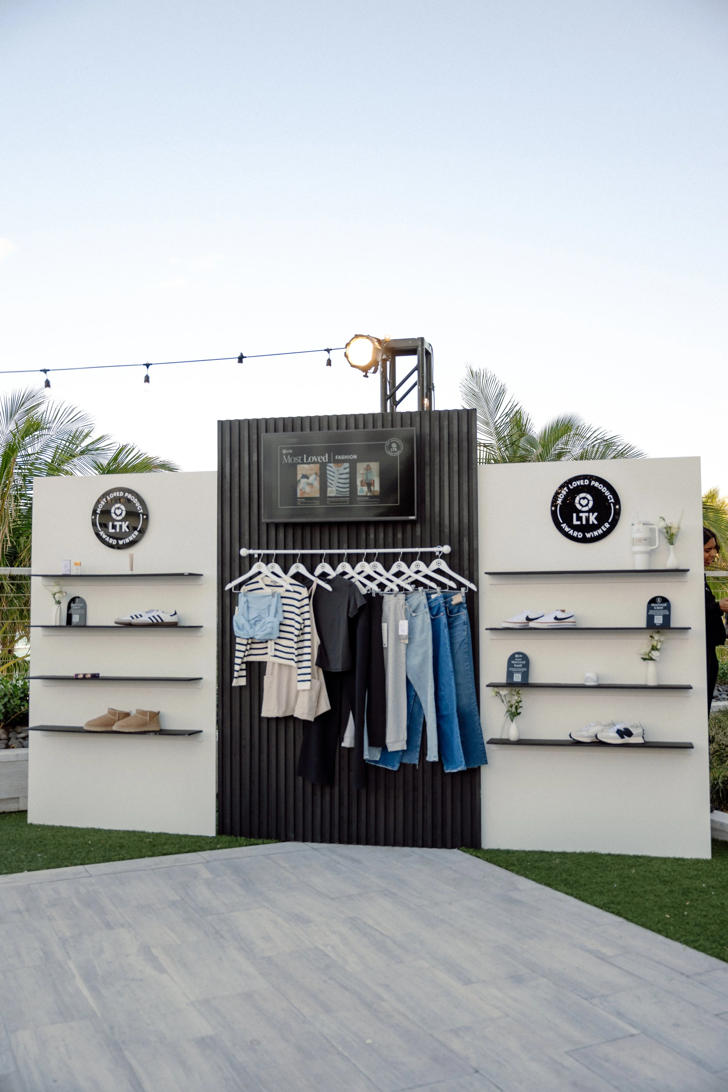 Outdoor clothing display with shelves holding shoes, bags, and accessories, against a white wall featuring round black logo signs and a black wooden panel with hanging clothes, set in a tropical environment with palm trees and string lights.