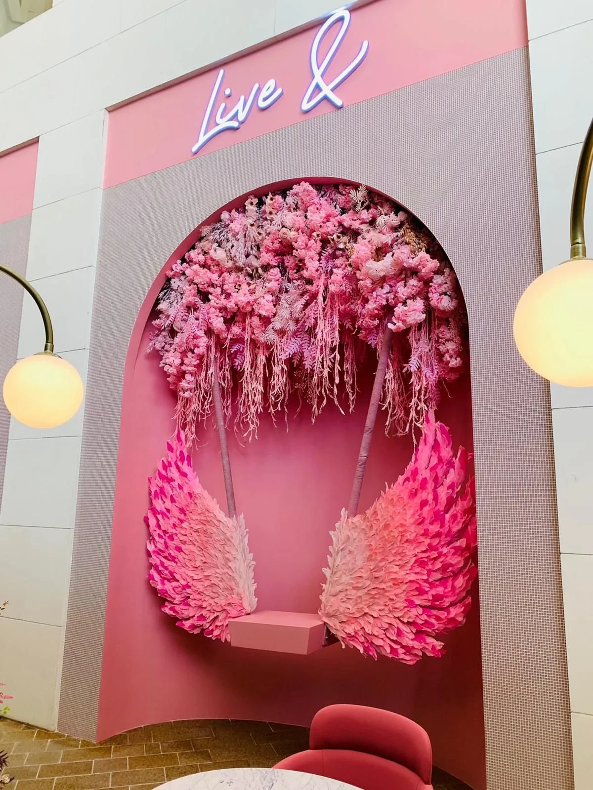 Decorative pink wall with white neon sign reading 'Live &', pink floral wall art resembling a swing with pink flowers and feather-like details, pink chair, and round wall sconces.