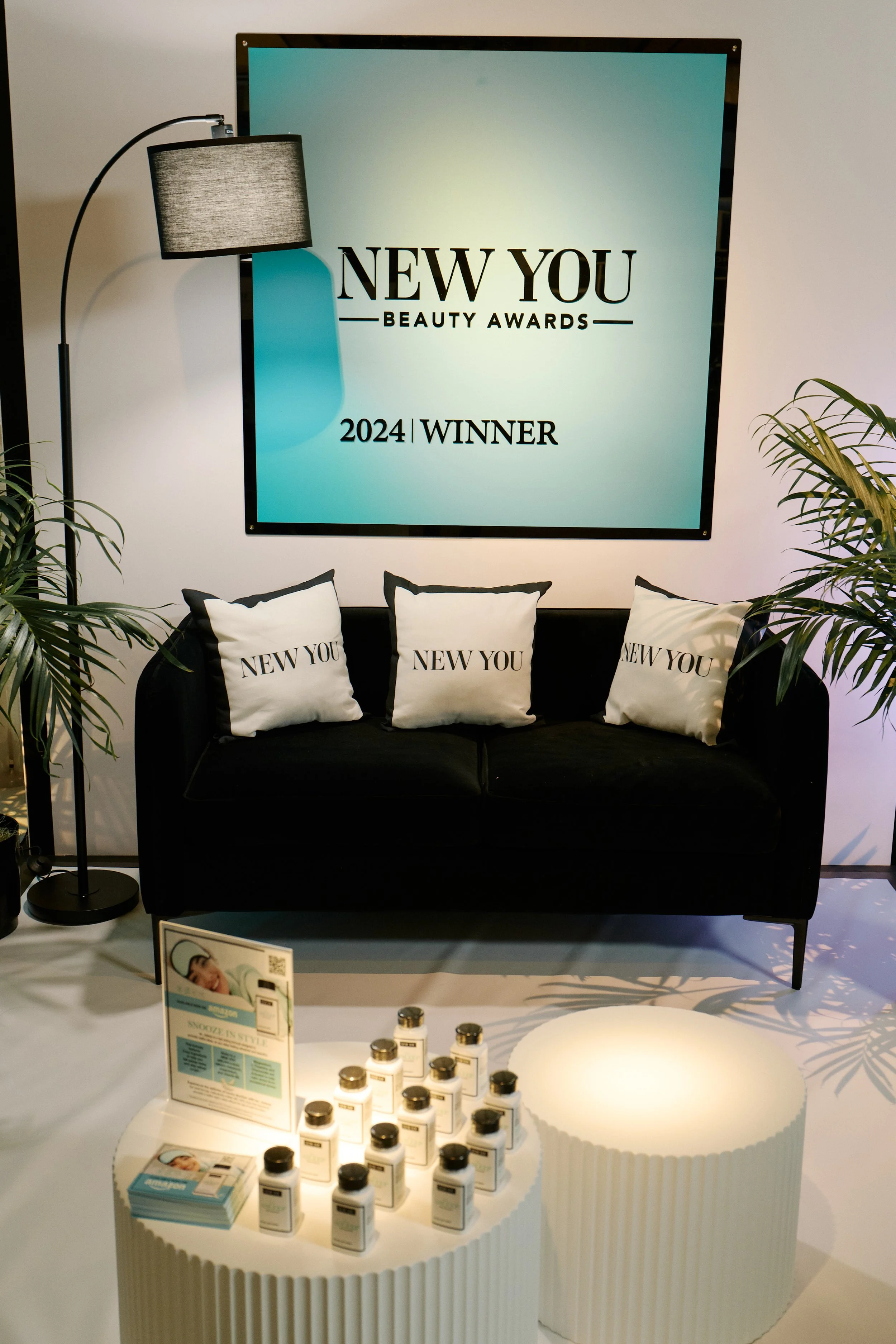 Display setup for New You Beauty Awards 2024 with a black sofa holding three pillows that have 'New You' written on them. There is a black floor lamp and a large digital screen in the background showing the event name and year. In the foreground, the