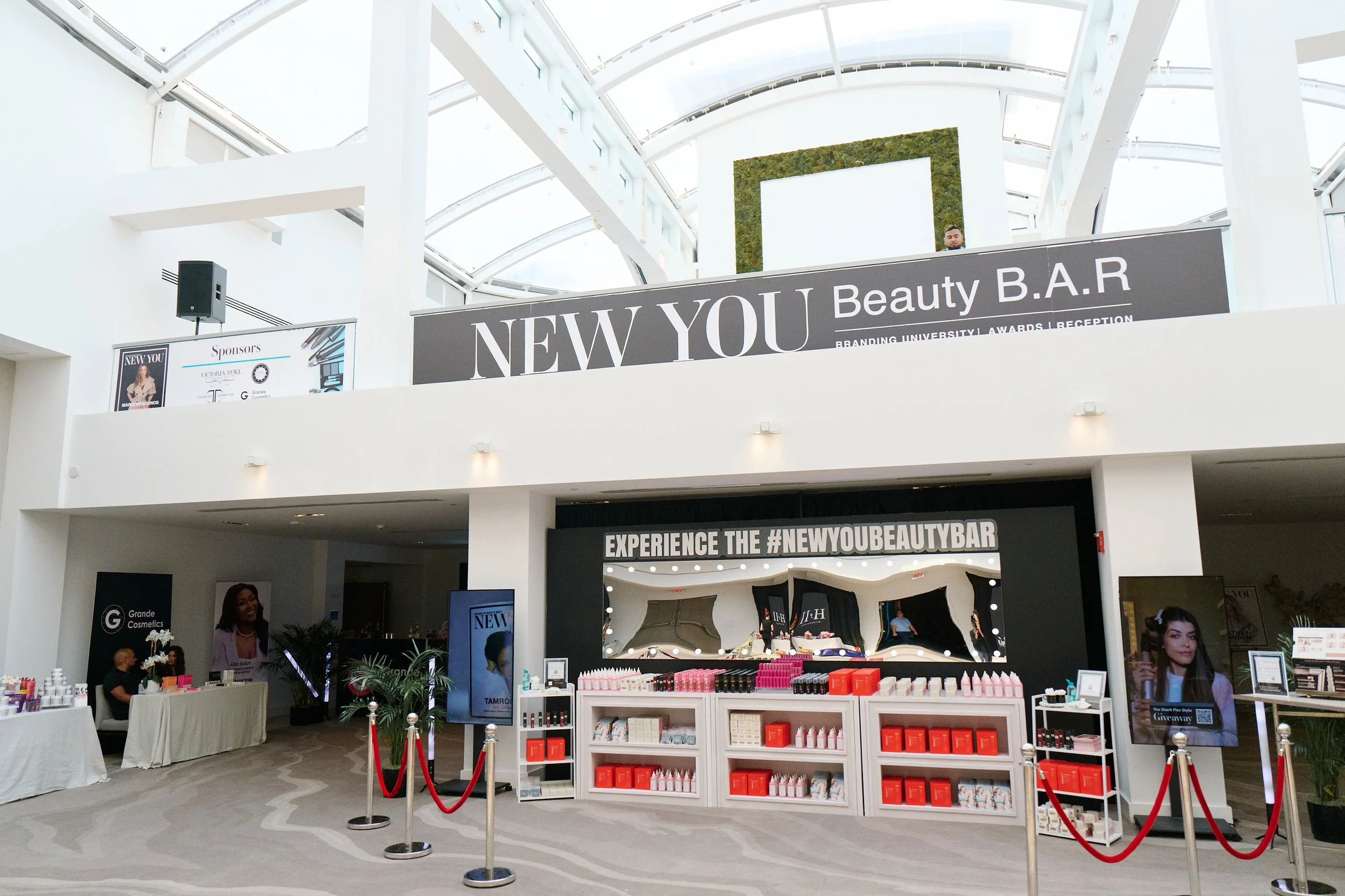 Interior of a shopping mall or convention center with a beauty bar named 'New You' on the second level, featuring beauty products and displays, with people working and browsing.