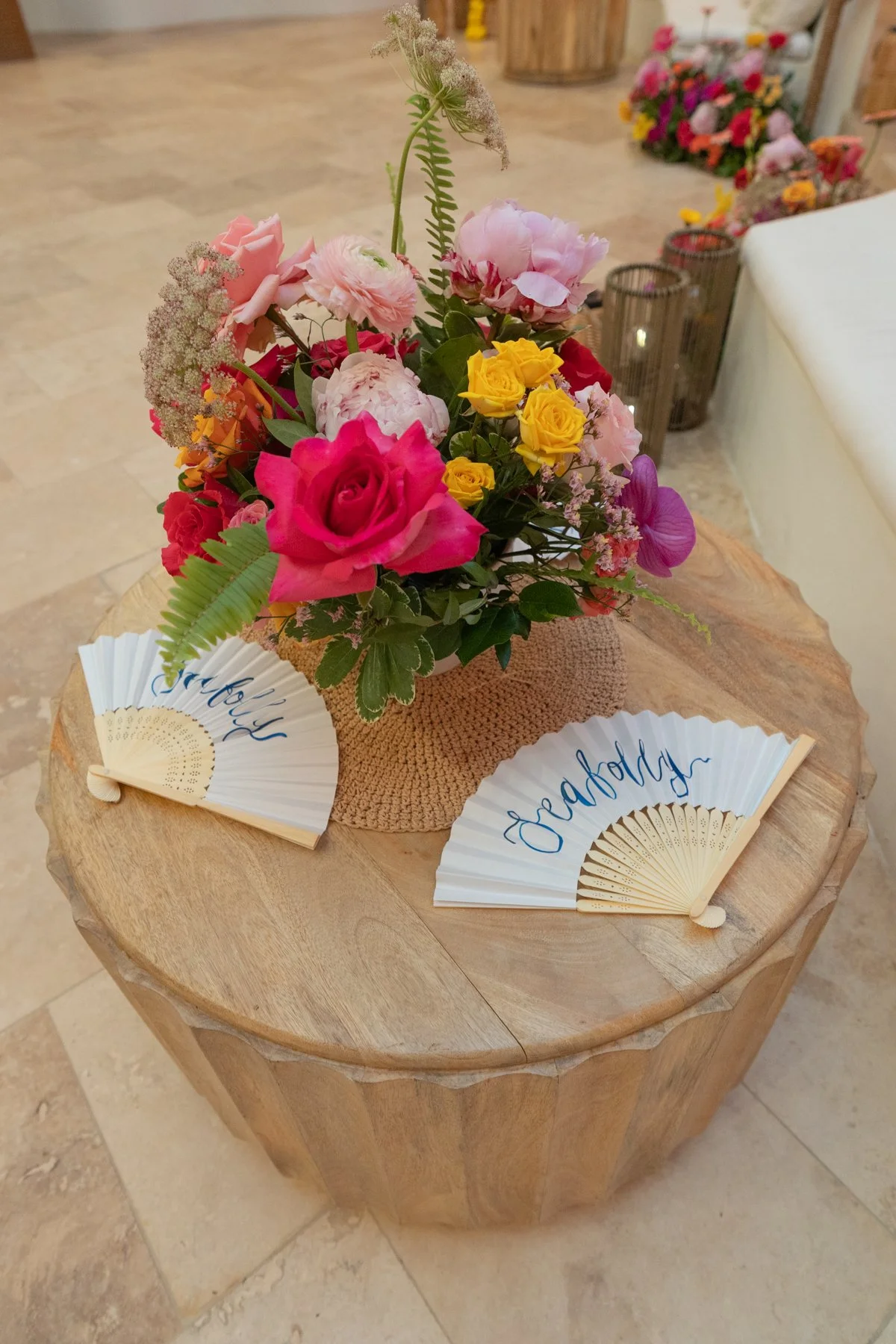 A floral arrangement of pink, yellow, and purple flowers in a woven basket on a wooden table, flanked by white paper fans with 'Hello' and 'Goodbye' written in blue.