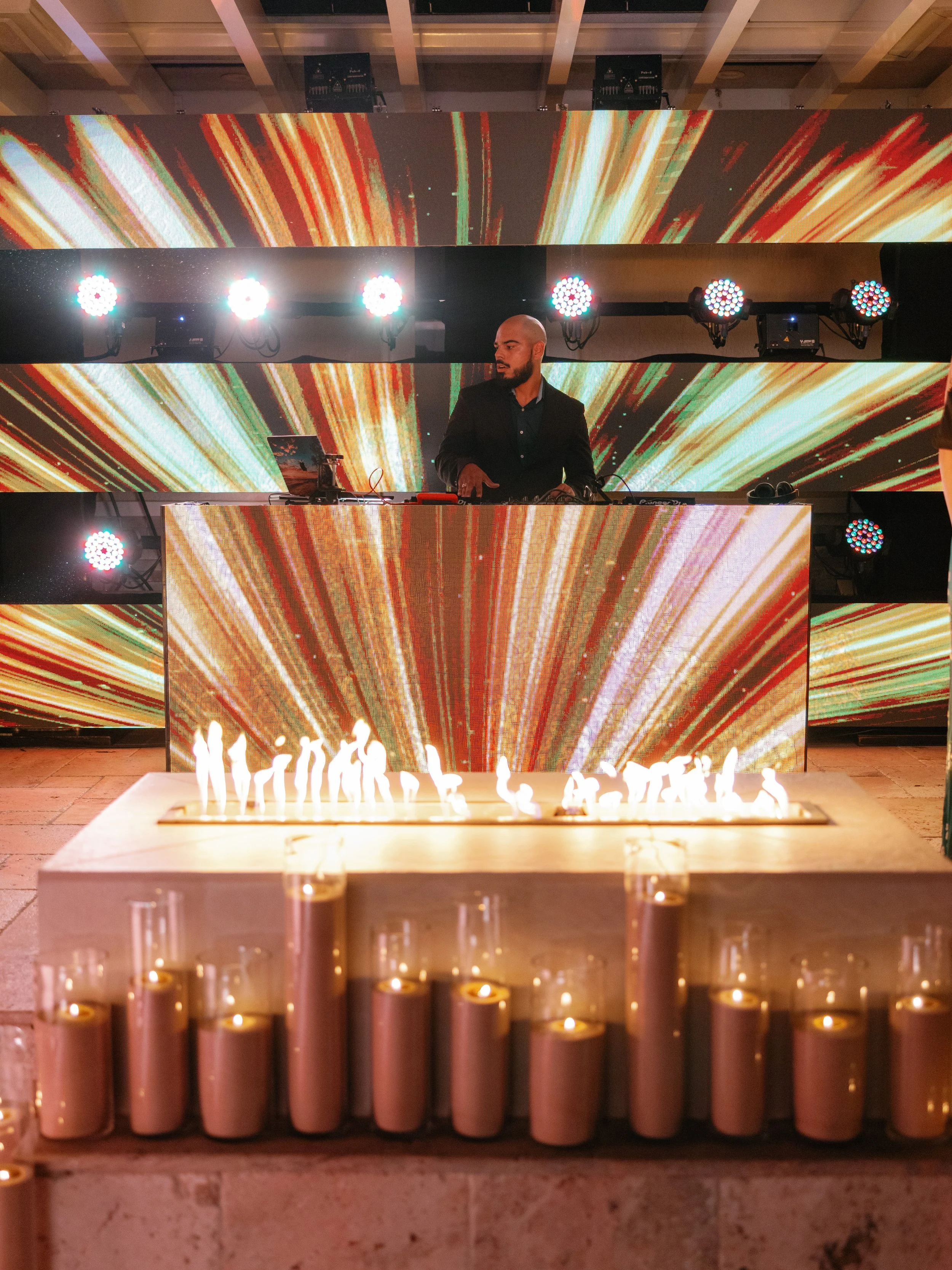 A man in a black suit standing behind a DJ booth with colorful digital screens in the background, decorated with candles in the foreground.