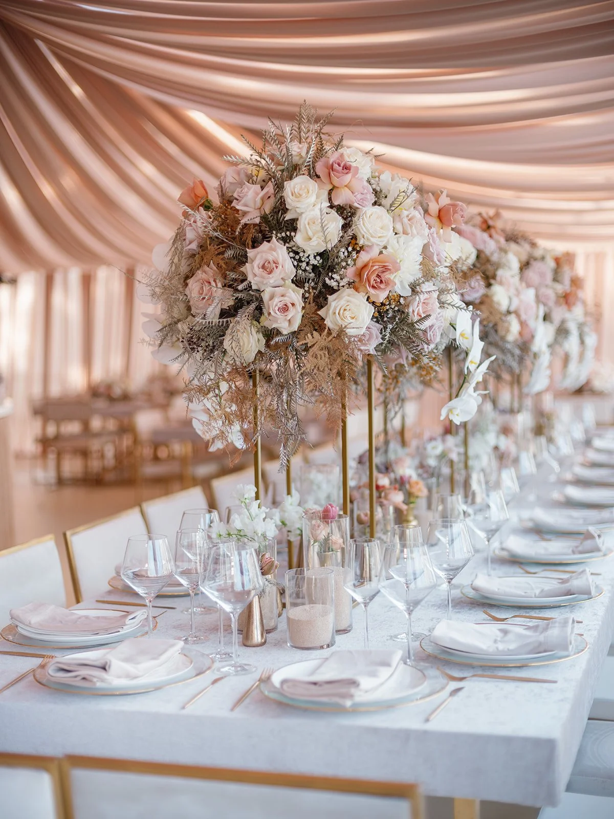 Elegant wedding table with tall floral centerpieces of pink, white roses, and greenery, set with white plates, glasses, and silverware under a soft pink draped ceiling.