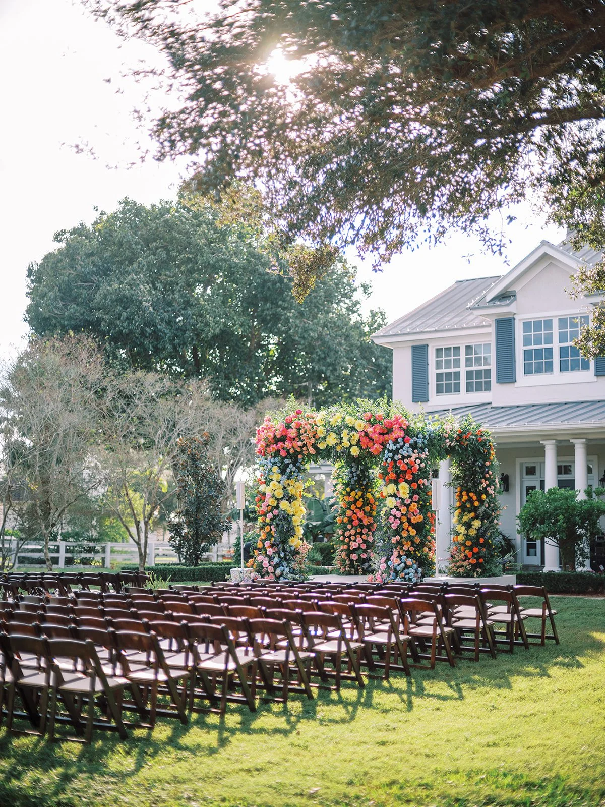 A wedding setup on the lawn in front of a white house with large windows. Rows of chairs face a decorated floral arch with pink, yellow, orange, purple, and white flowers. The scene is illuminated by sunlight through trees overhead.