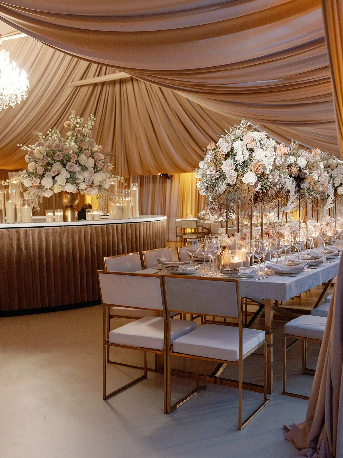 Elegant banquet setup with a long table decorated with large floral centerpieces, surrounded by chairs with white cushions, set for a formal dinner in a decorated venue with draped fabric ceilings and candles.