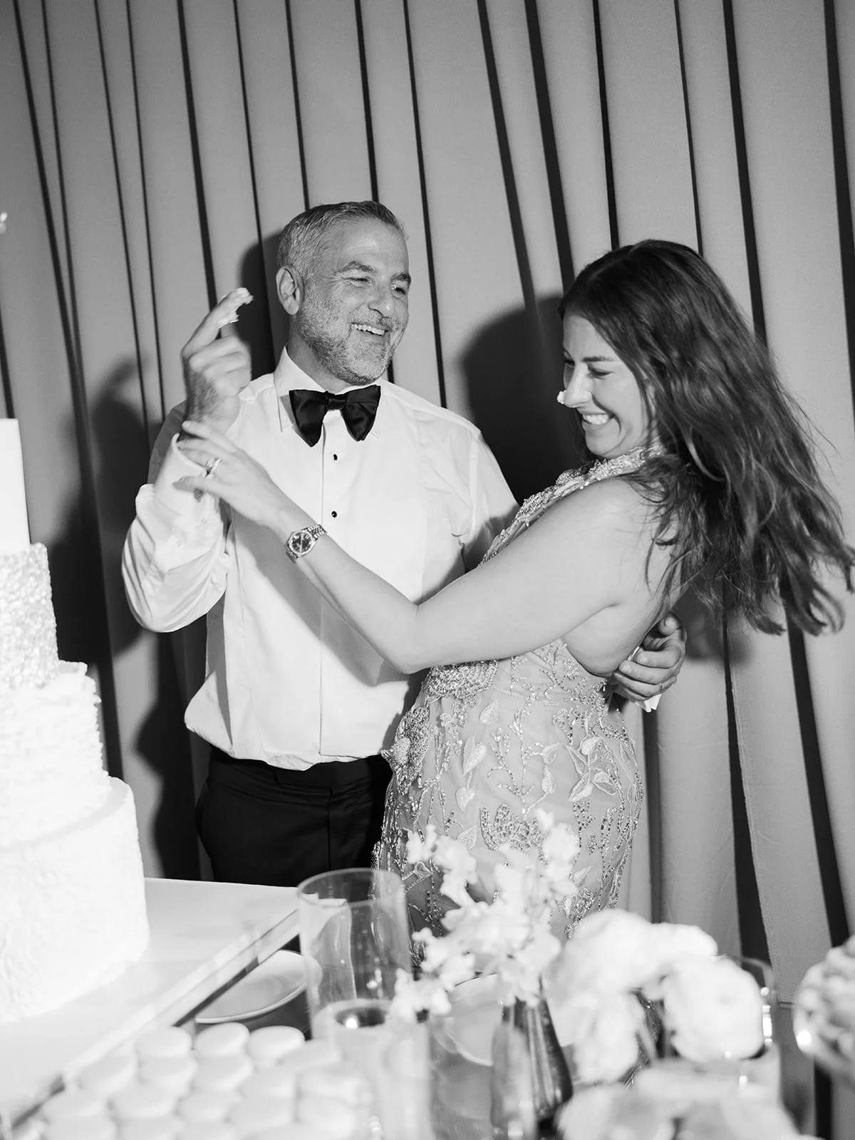 A black and white photo of a man and woman dancing at a celebration, possibly a wedding, with a decorated table and cake in the foreground.