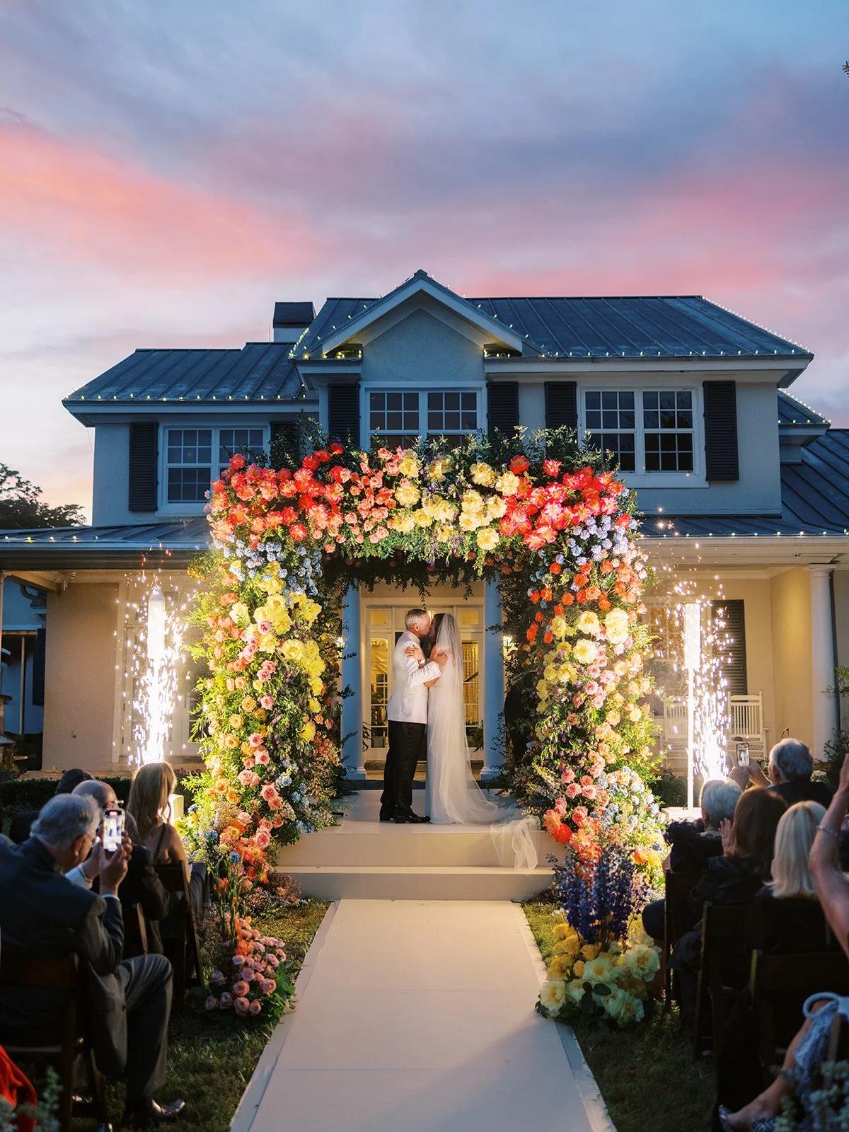 A wedding ceremony taking place outdoors in front of a large house, with a floral arch and fireworks. The couple is kissing under the arch, surrounded by guests.