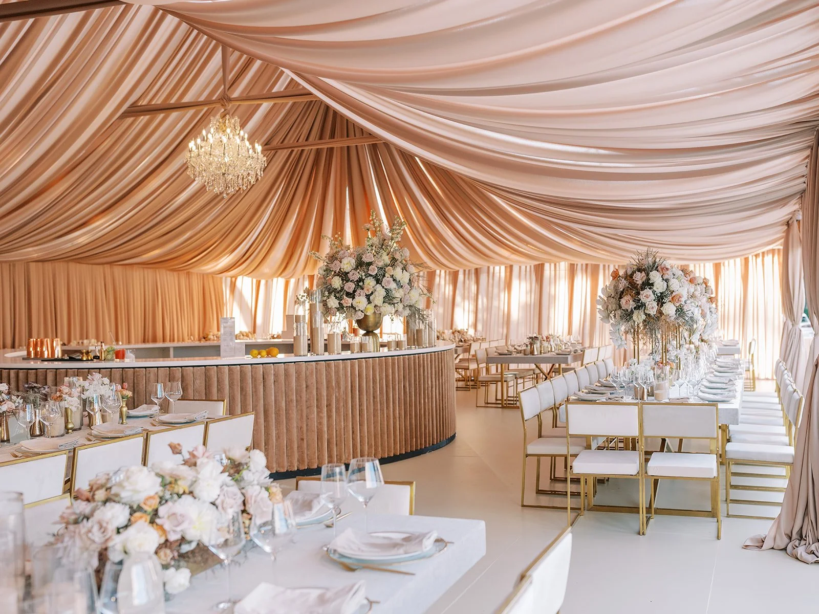 Elegant event space with draped peach-colored fabric ceiling, large floral centerpieces on tables, white table settings, and a bar in the background.